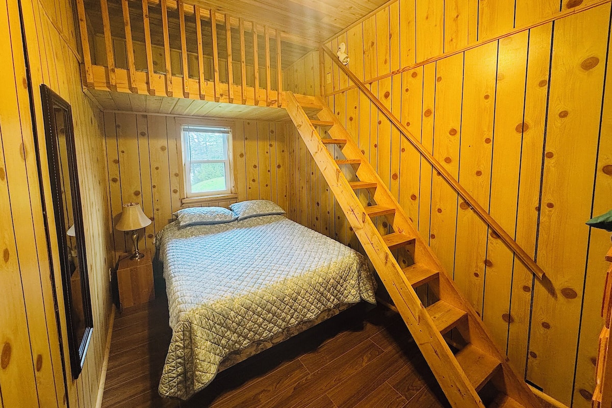 A wooden cabin bedroom with a bed, a nightstand, a lamp, a window, and a wooden staircase.