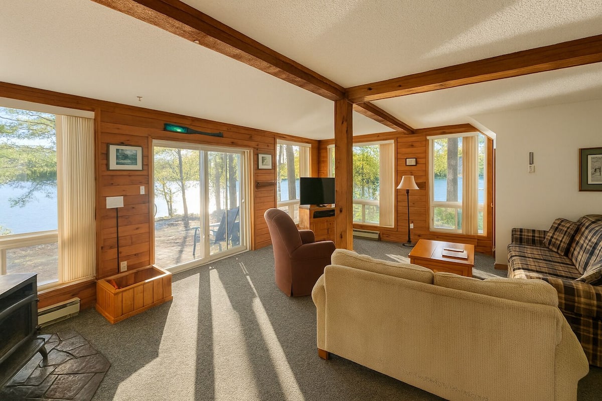 A cozy cabin interior with a fireplace, a television, and a large window overlooking a lake.