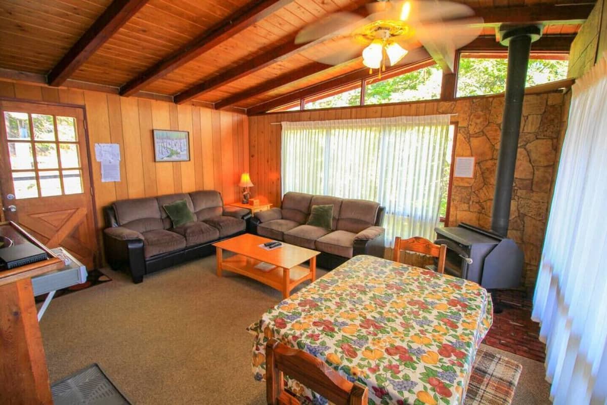 A cozy cabin interior with a wooden ceiling, a stone wall, a fireplace, a sofa set, a dining table, and a bed with a floral bedspread.