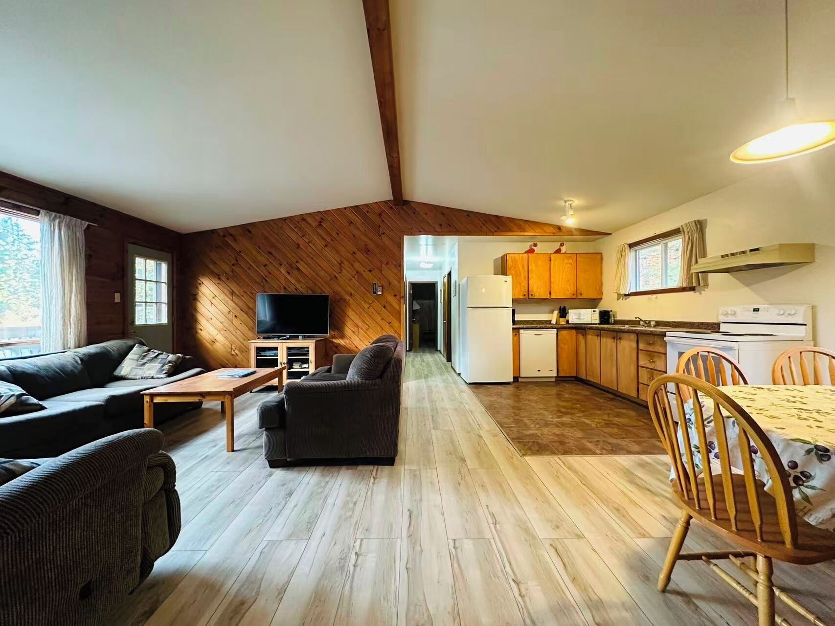 Un grand salon comprenant un canapé, un téléviseur et une table à manger avec des chaises. La cuisine est visible et est équipée de meubles en bois et d’un réfrigérateur.