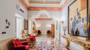 Un amplio pasillo con piso a cuadros, sillones tapizados en rojo y un cuadro grande y ornamentado.