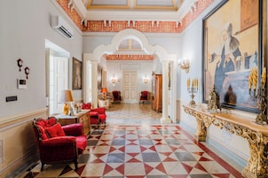 Un amplio pasillo con piso a cuadros, sillones tapizados en rojo y un cuadro grande y ornamentado.