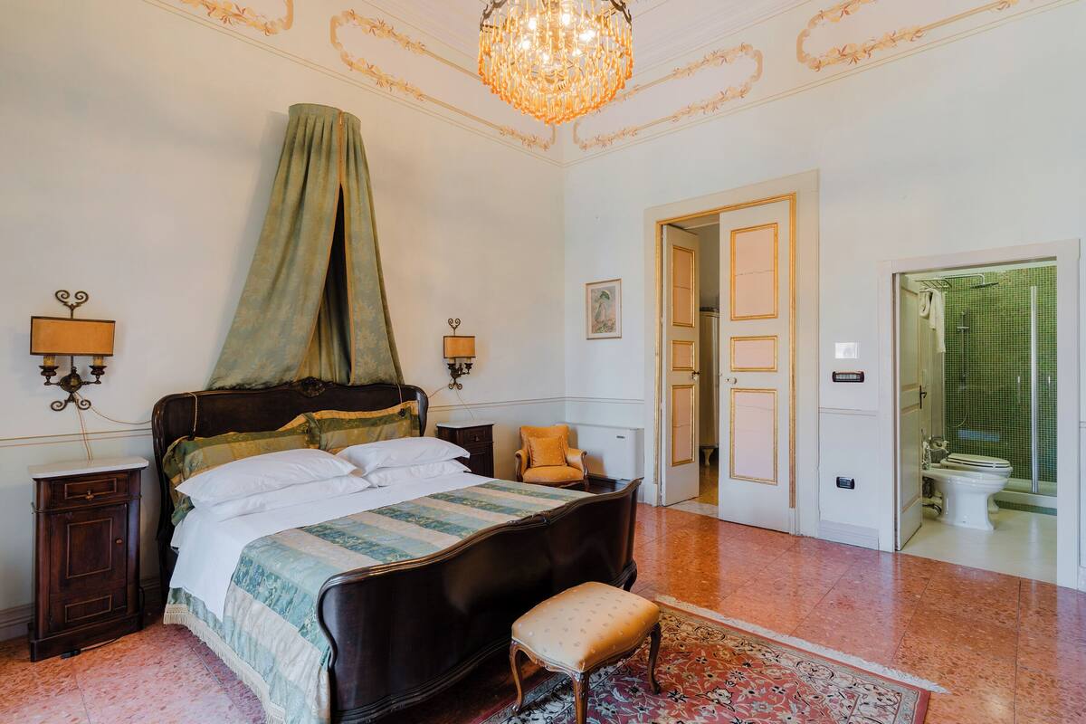 Un dormitorio con una cama grande, una silla, una mesita de noche, una araña y un baño visible a través de una puerta abierta.