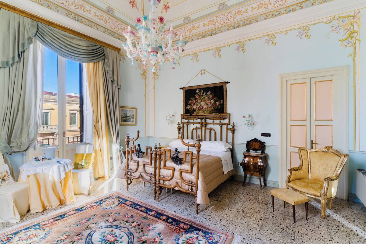 Una habitación con cama, una silla, una mesa, un candelabro y una ventana con cortinas.