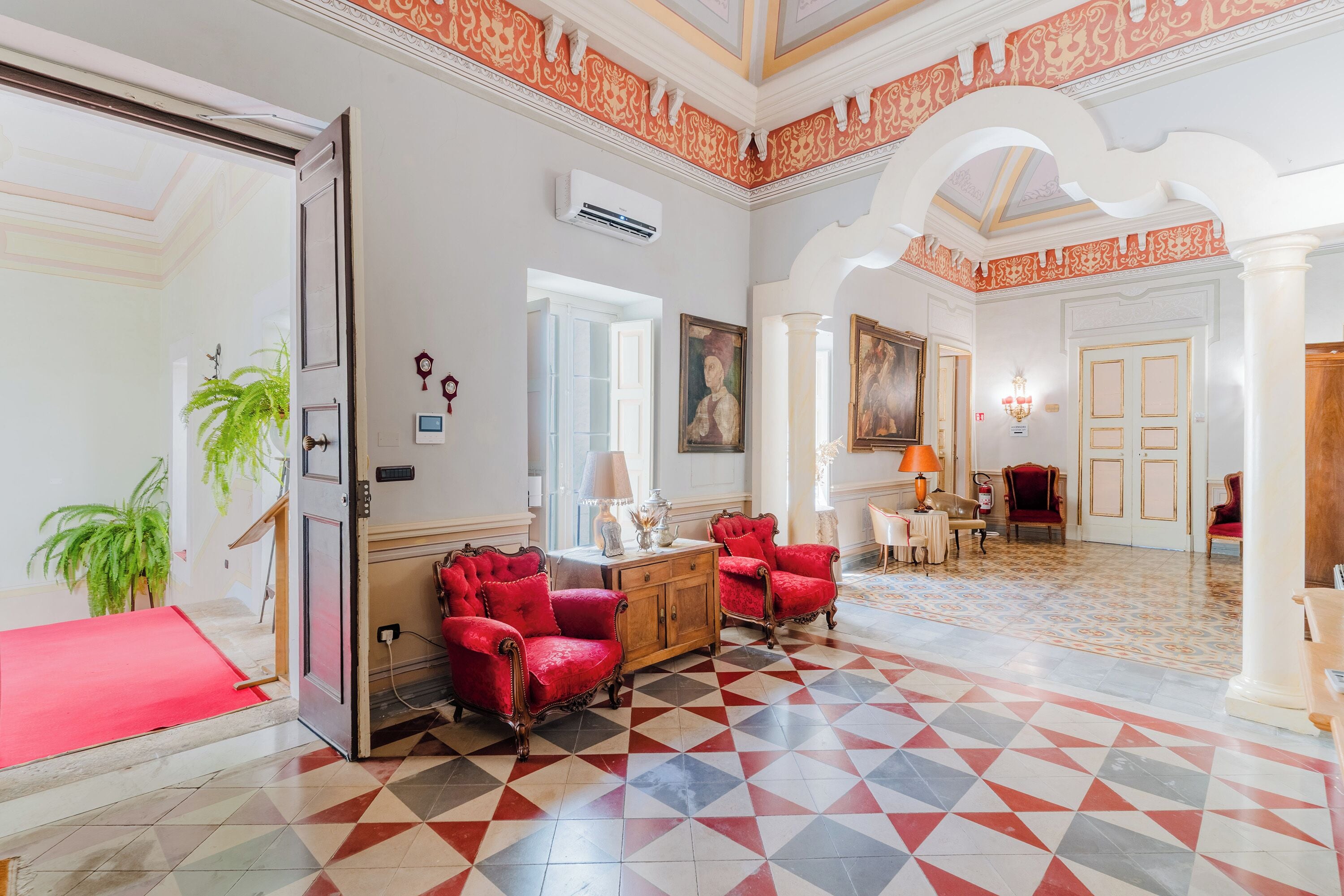 Un hall d’hôtel avec un comptoir d’enregistrement, des fauteuils rouges, un sol à motifs et un grand tableau accroché au mur.