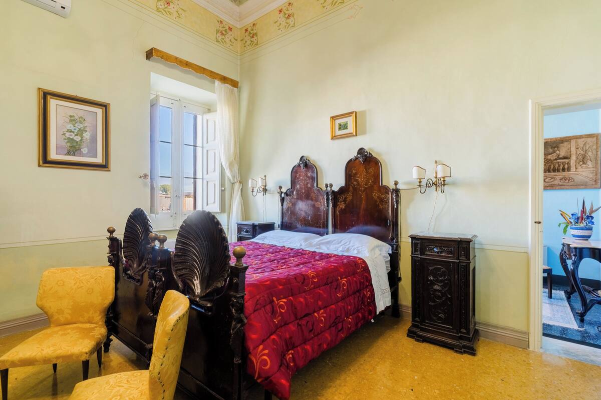 Un dormitorio con una cama grande, dos sillones, una mesita de noche y un ventanal con cortinas.
