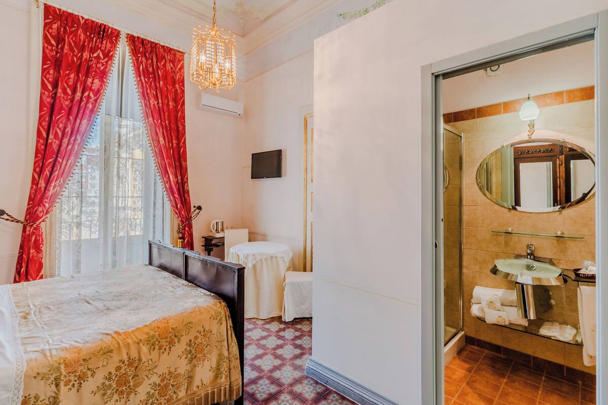 Habitación de hotel con cama, una mesita, una silla, televisor y baño con lavabo, espejo y toallero.