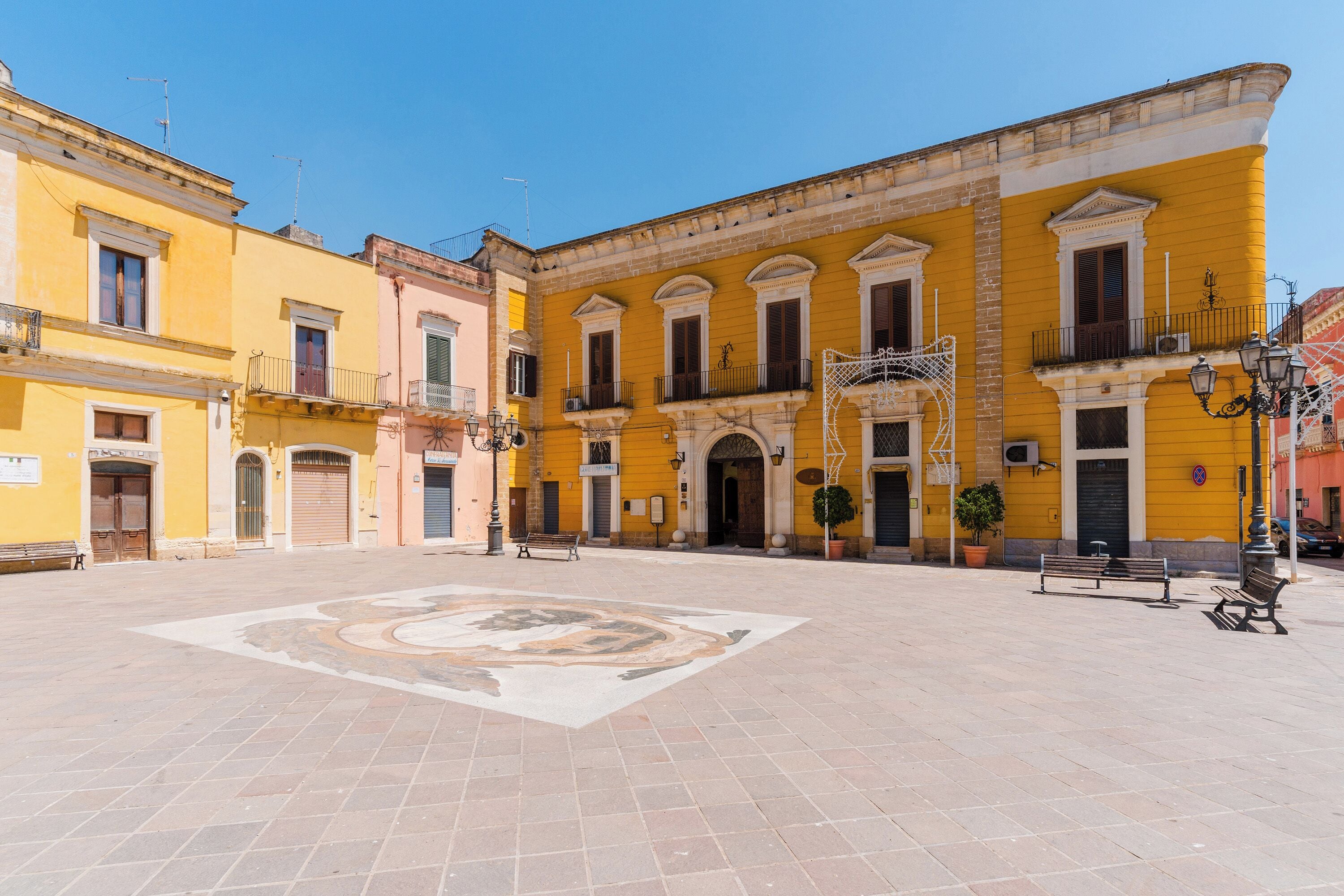 A spacious courtyard with a central mosaic, surrounded by yellow and pink buildings with balconies and arched doorways.