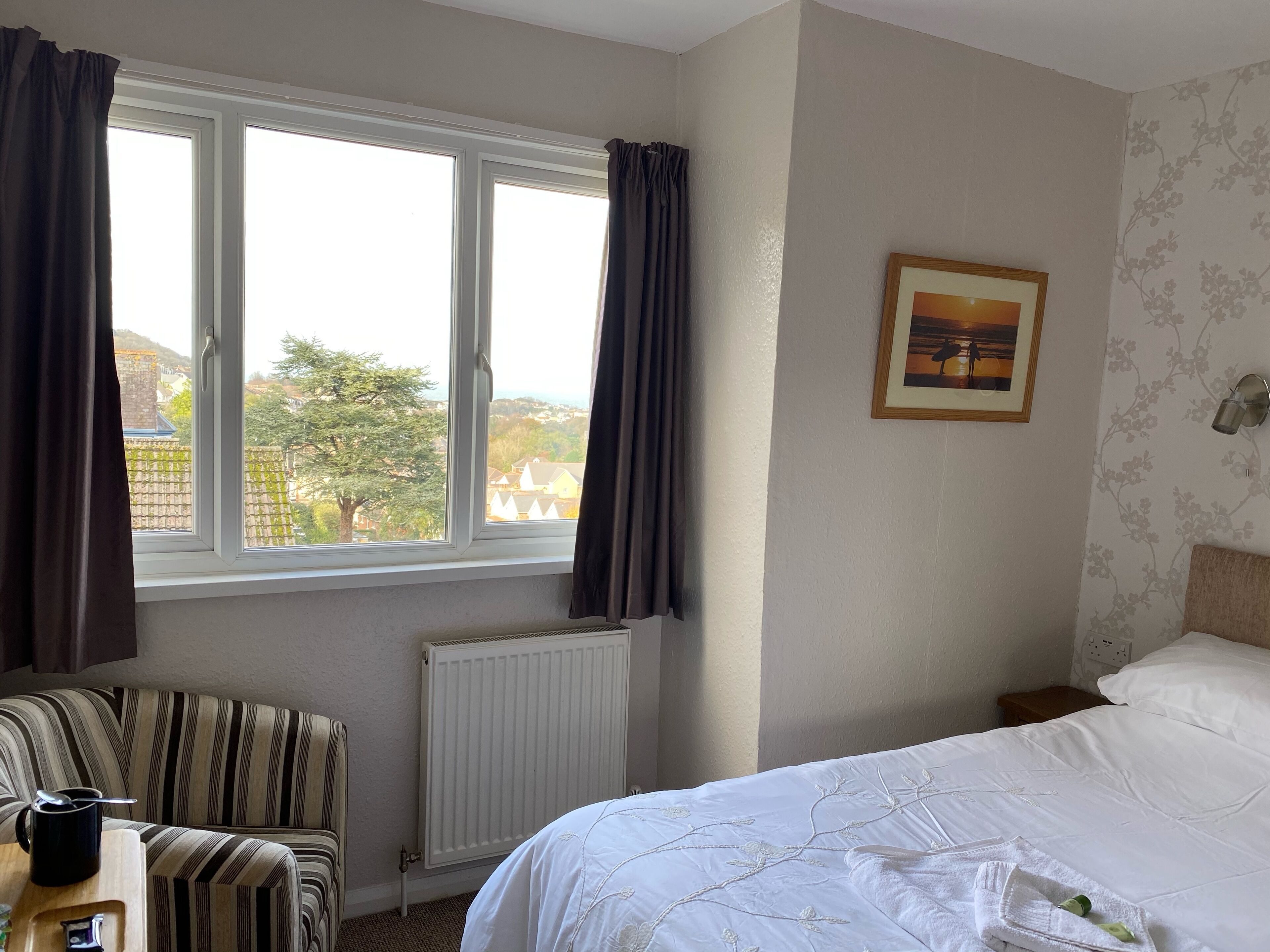 Una habitación de hotel con una cama, un sillón a rayas, un radiador y una ventana con cortinas.