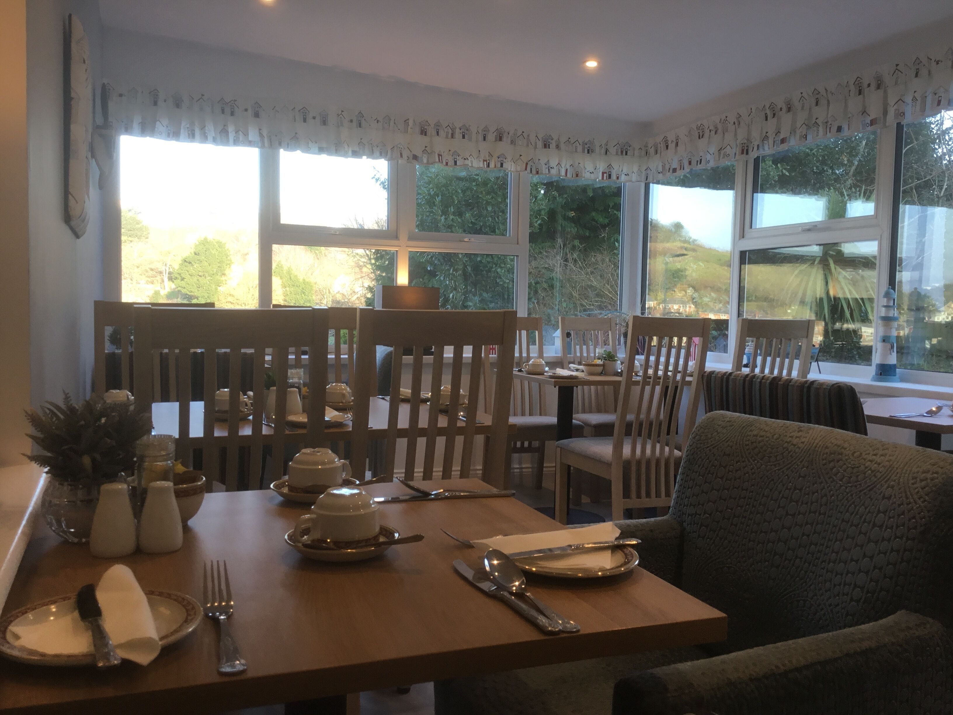 A dining area with wooden chairs and tables, a view of greenery outside, and a decorative house pattern on the window valance.