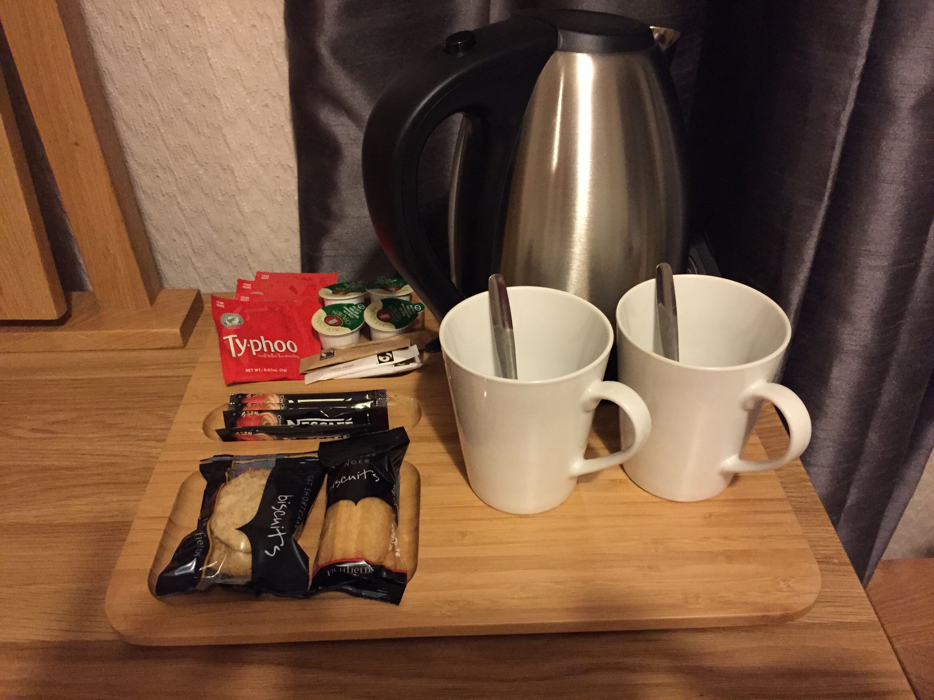 A wooden tray with a coffee pot, two white mugs, a packet of biscuits, and a bag of Tygphoo tea.
