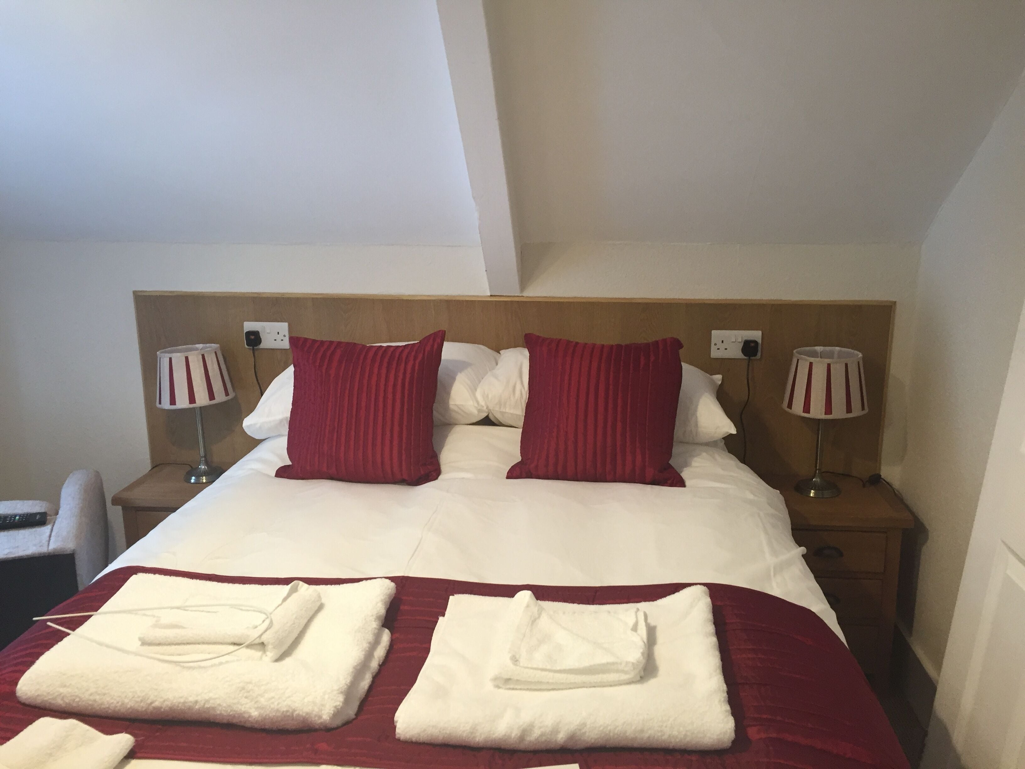 A hotel room with a large bed, two red pillows, white bedding, two bedside lamps, and two folded towels on the bed.