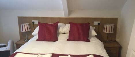 A hotel room with a large bed, two red pillows, white bedding, two bedside lamps, and two folded towels on the bed.