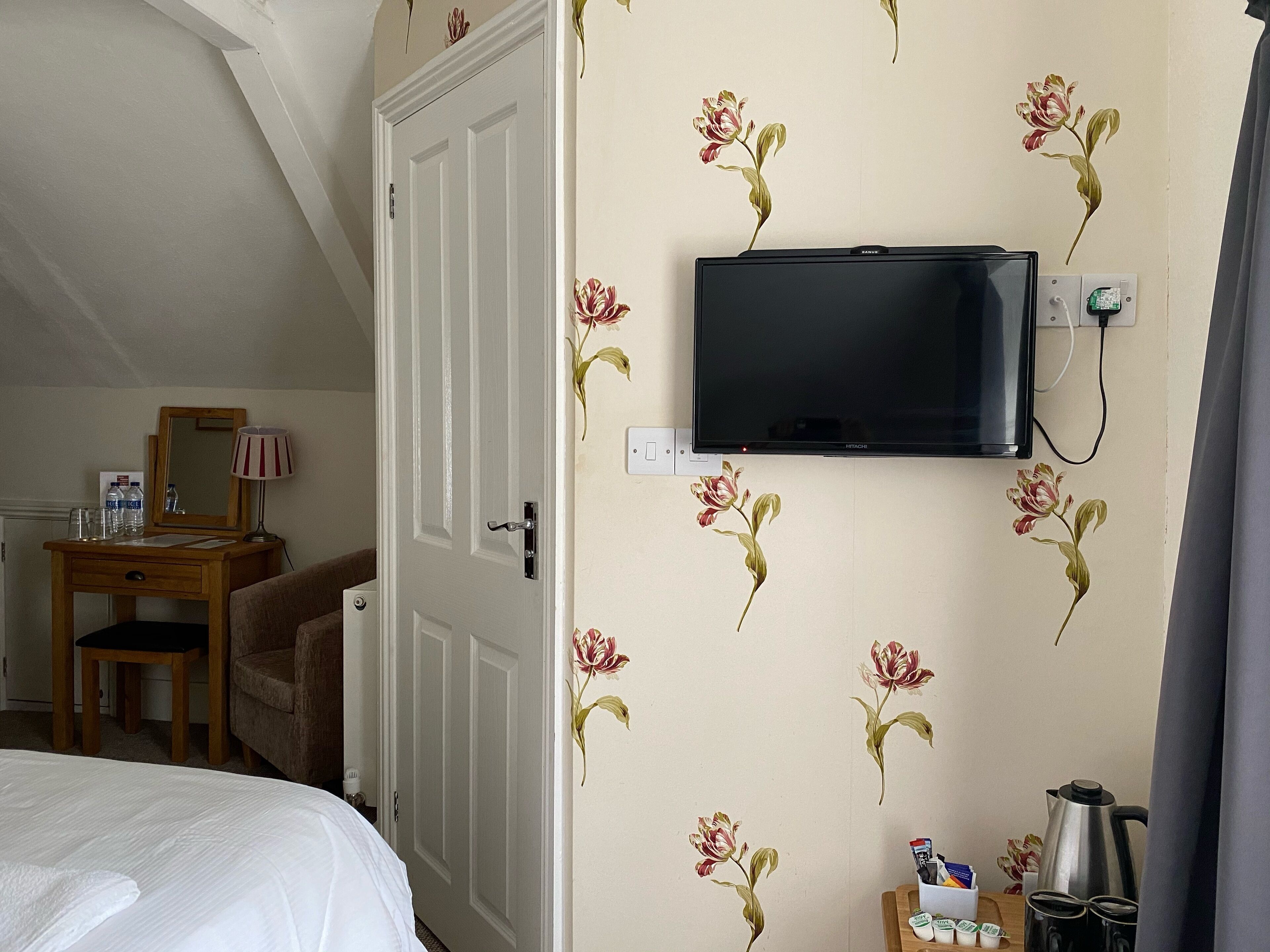 Una habitación de hotel con una cama, un escritorio con lámpara, un televisor montado en la pared y una cafetera sobre la mesita de noche.
