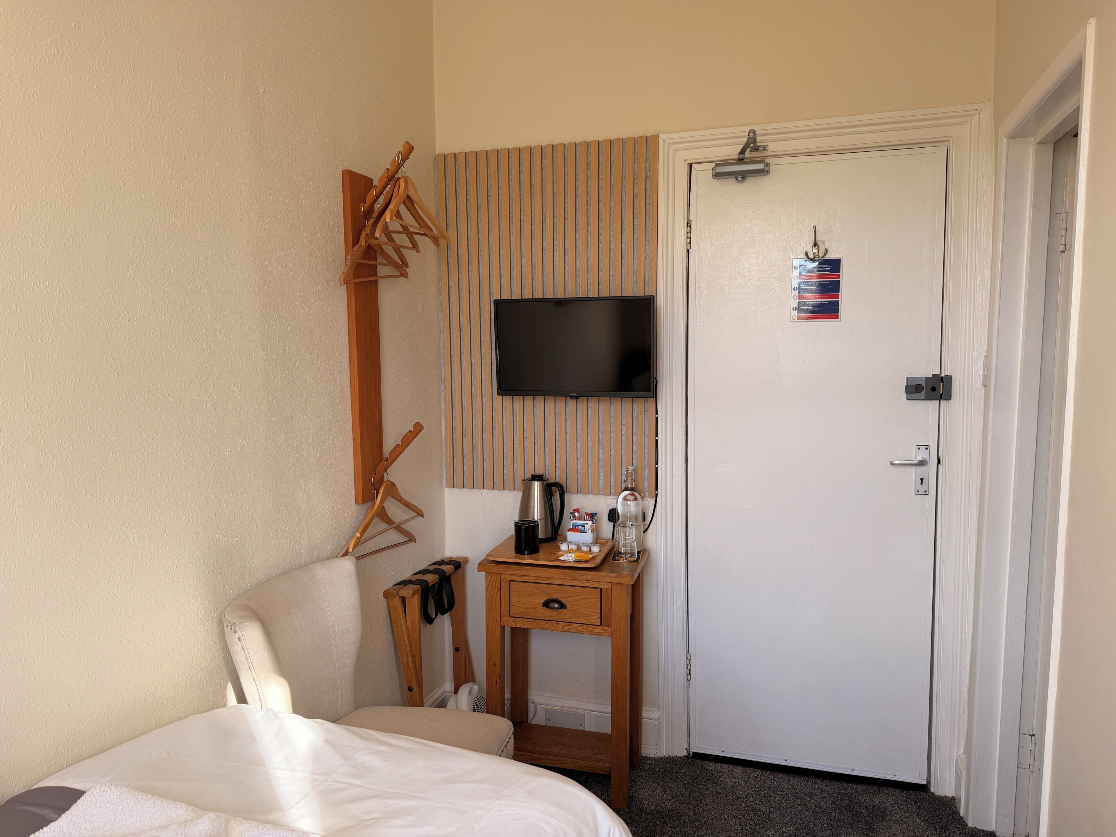 A hotel room with a bed, a wooden chair, a TV mounted on the wall, a small wooden table with a kettle and cups, and a door with a lock.