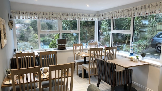 A dining area with wooden furniture, large windows with house-patterned curtains, and a view of greenery outside. - Cairn House (Ilfracombe)