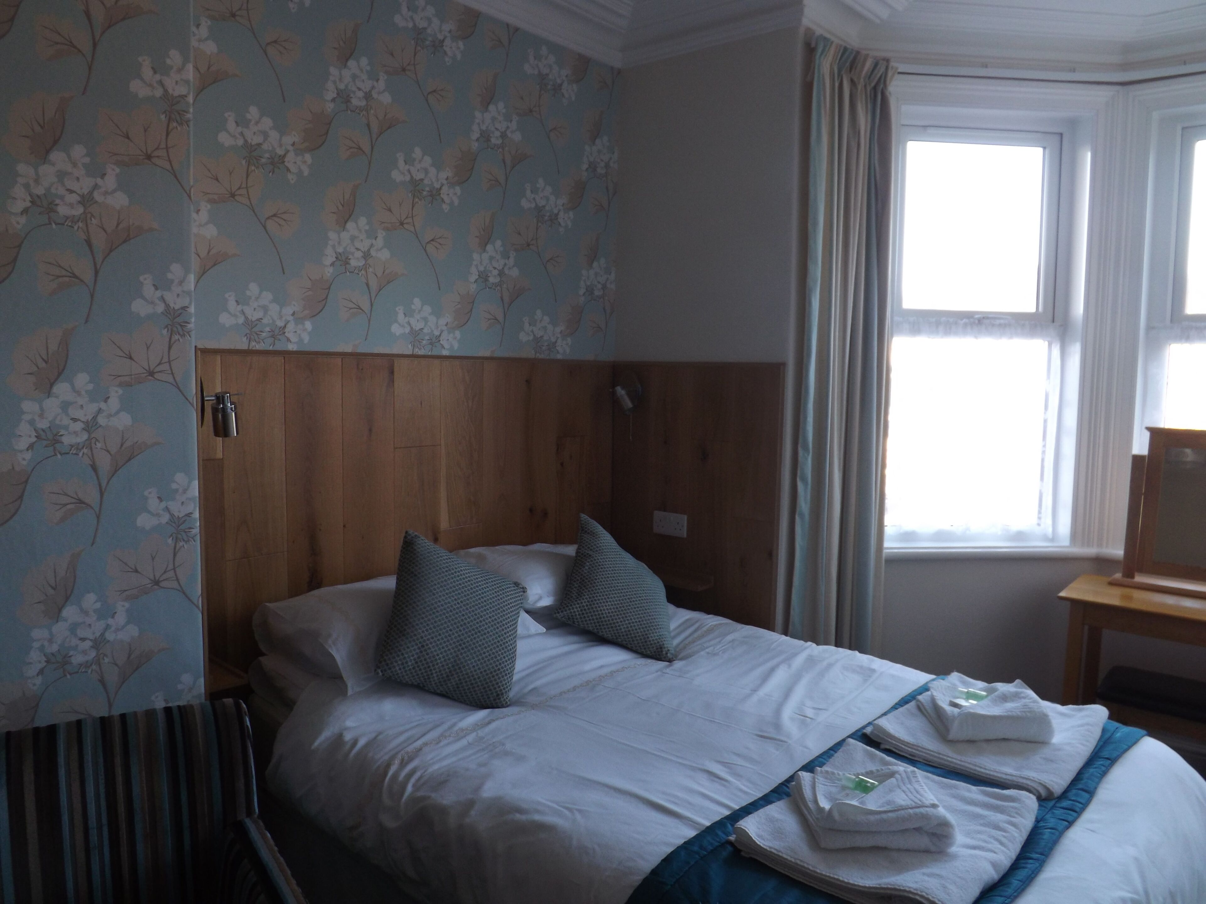 Habitación de hotel con cama, dos almohadas, una colcha azul y una ventana con cortinas.