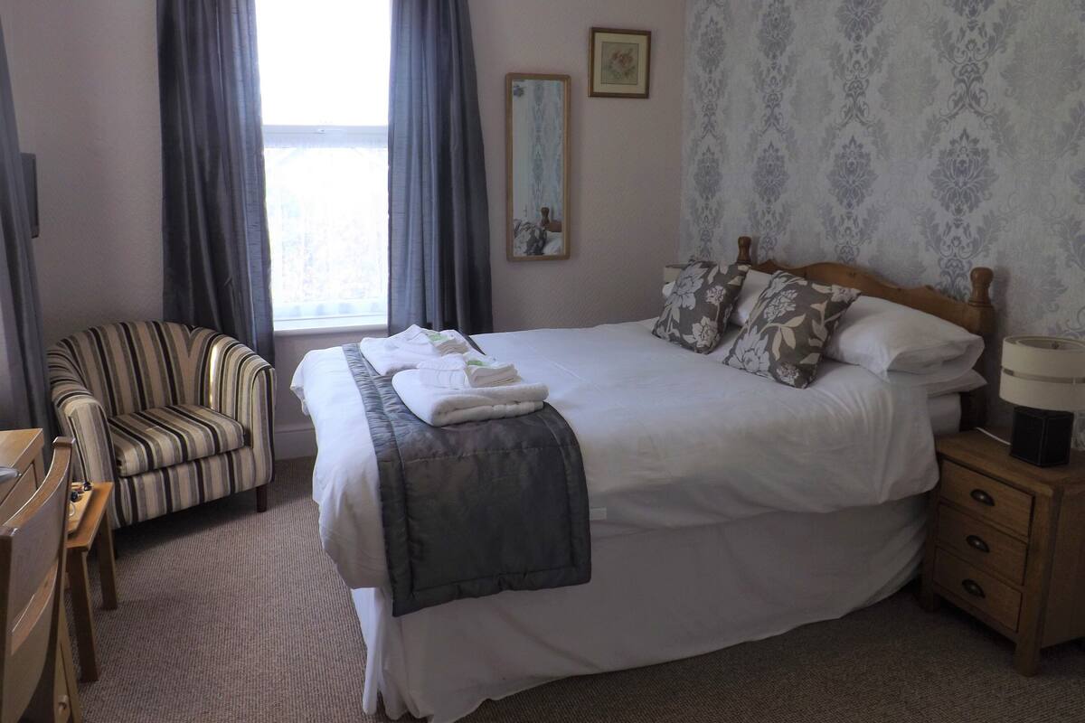 A neatly arranged hotel room with a large bed, a striped armchair, a wooden nightstand, and a window with curtains.