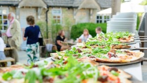 A dining area with tables set for meals, featuring pizzas with toppings and a rustic stone building in the background. - Old Stocks Inn (Cheltenham)