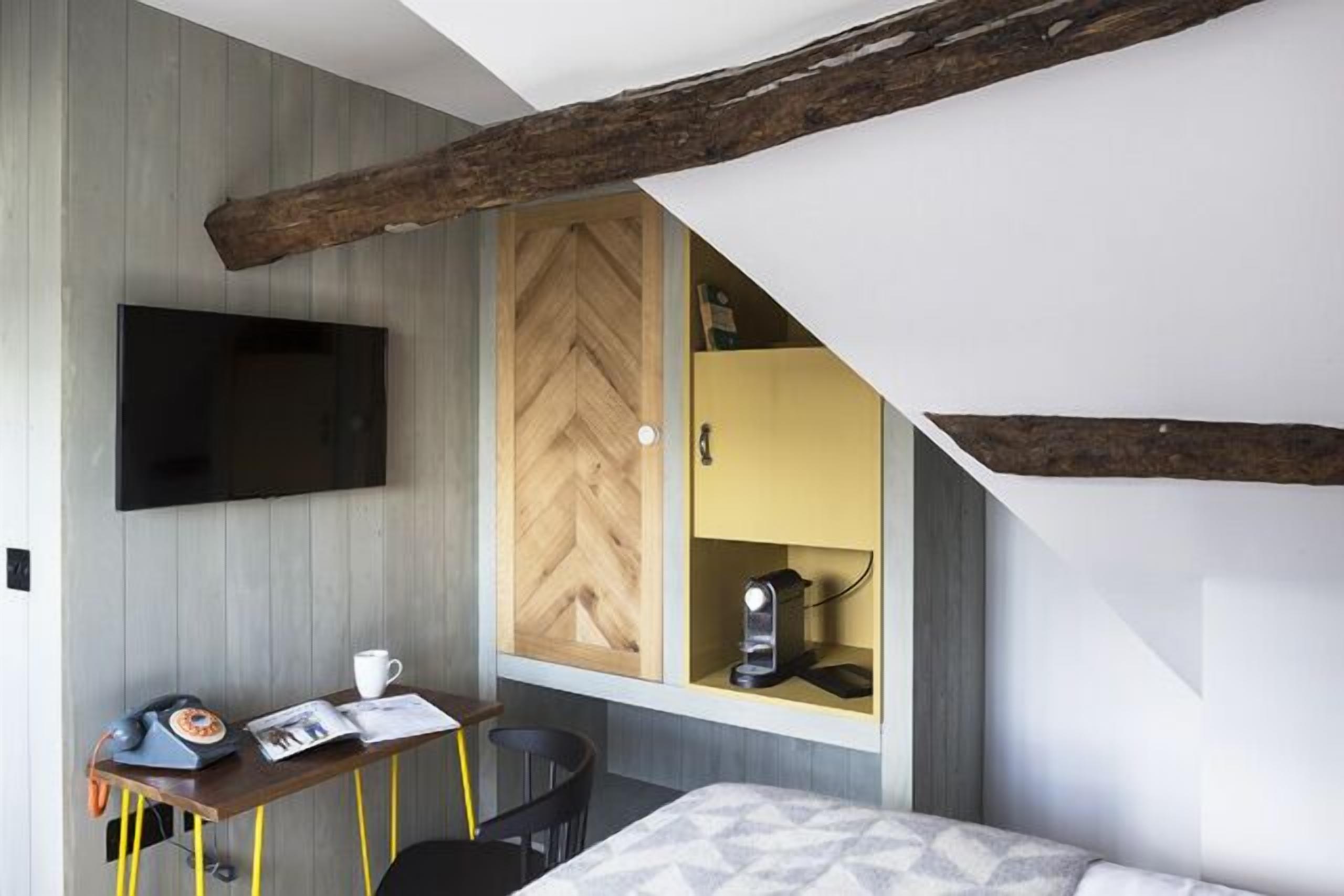 Un dormitorio con cama, escritorio, teléfono, taza y revista. Hay un televisor montado en la pared y una ventana con puerta de madera.