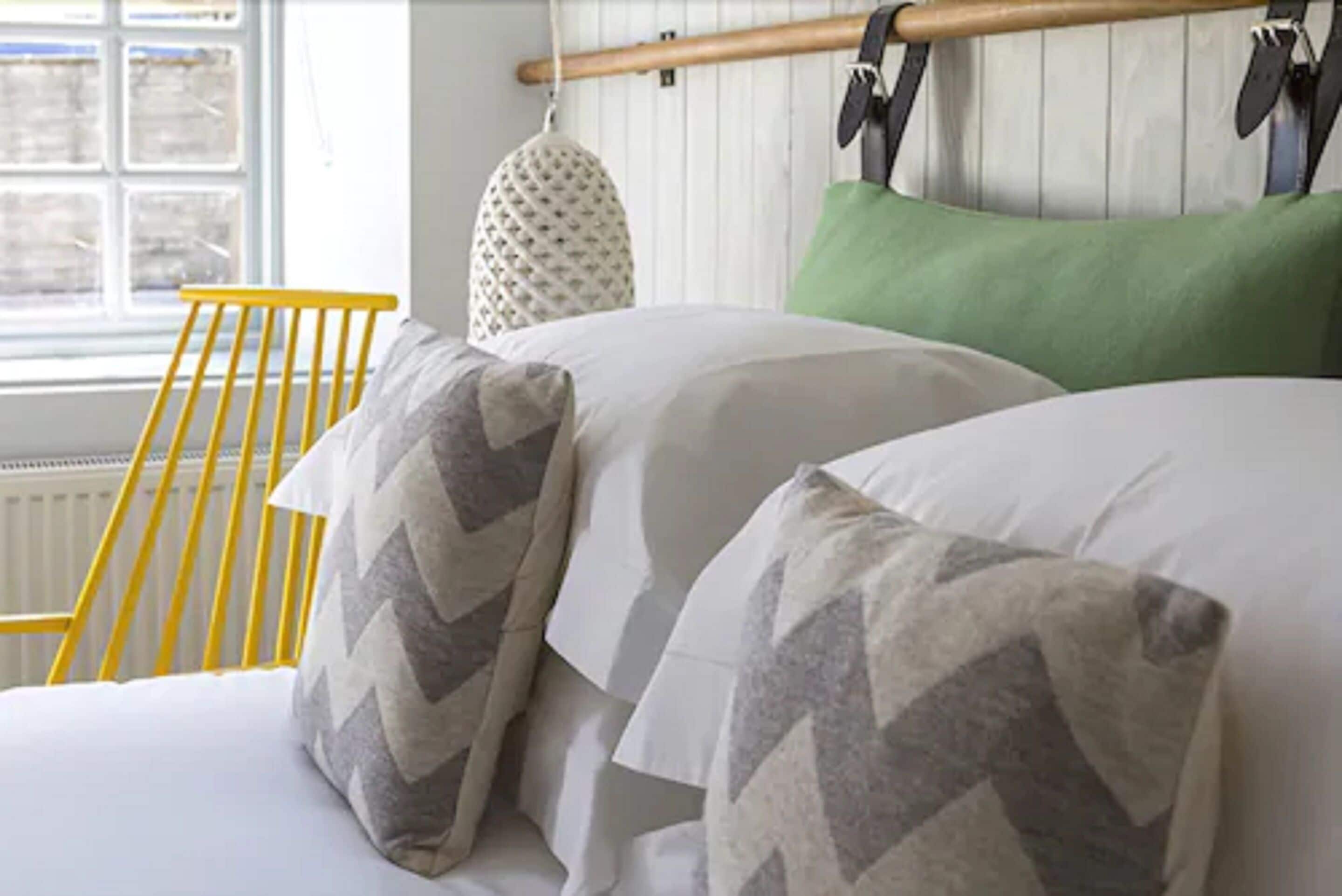 A bed with white and gray patterned pillows, a green pillow, and a yellow chair in a room with a window and a radiator.