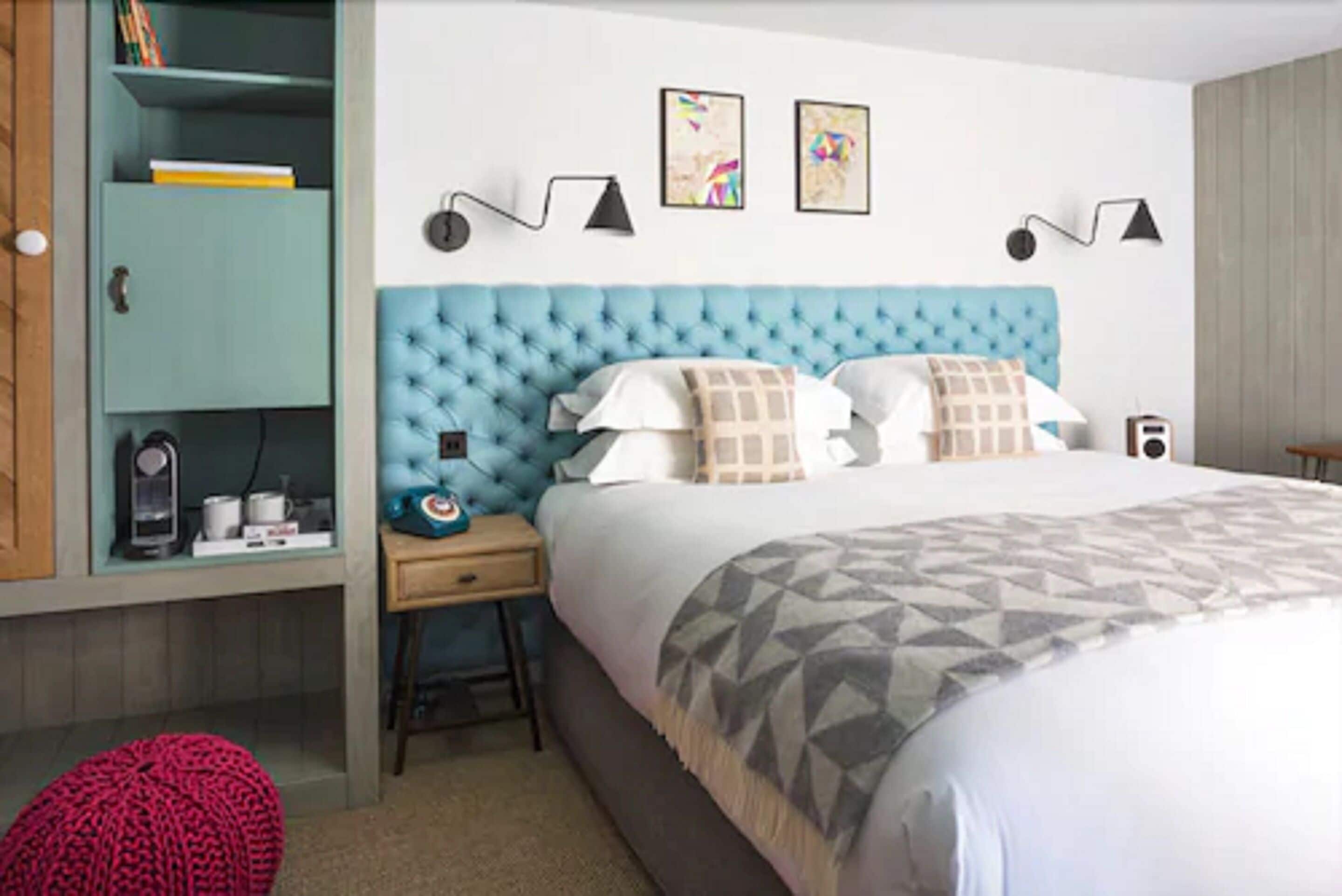Une chambre à coucher avec une tête de lit capitonnée, un lit avec une literie à motifs, une table de chevet avec une lampe, une armoire et un pouf rouge.