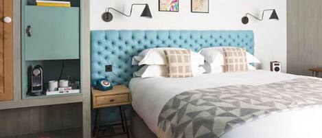 Une chambre à coucher avec une tête de lit capitonnée, un lit avec une literie à motifs, une table de chevet avec une lampe, une armoire et un pouf rouge.