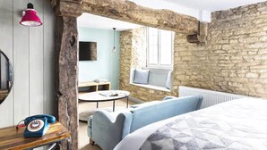 A rustic room with exposed wooden beams, a stone wall, a round table, a blue sofa, and a bed with a patterned cover. - Old Stocks Inn (Cheltenham)