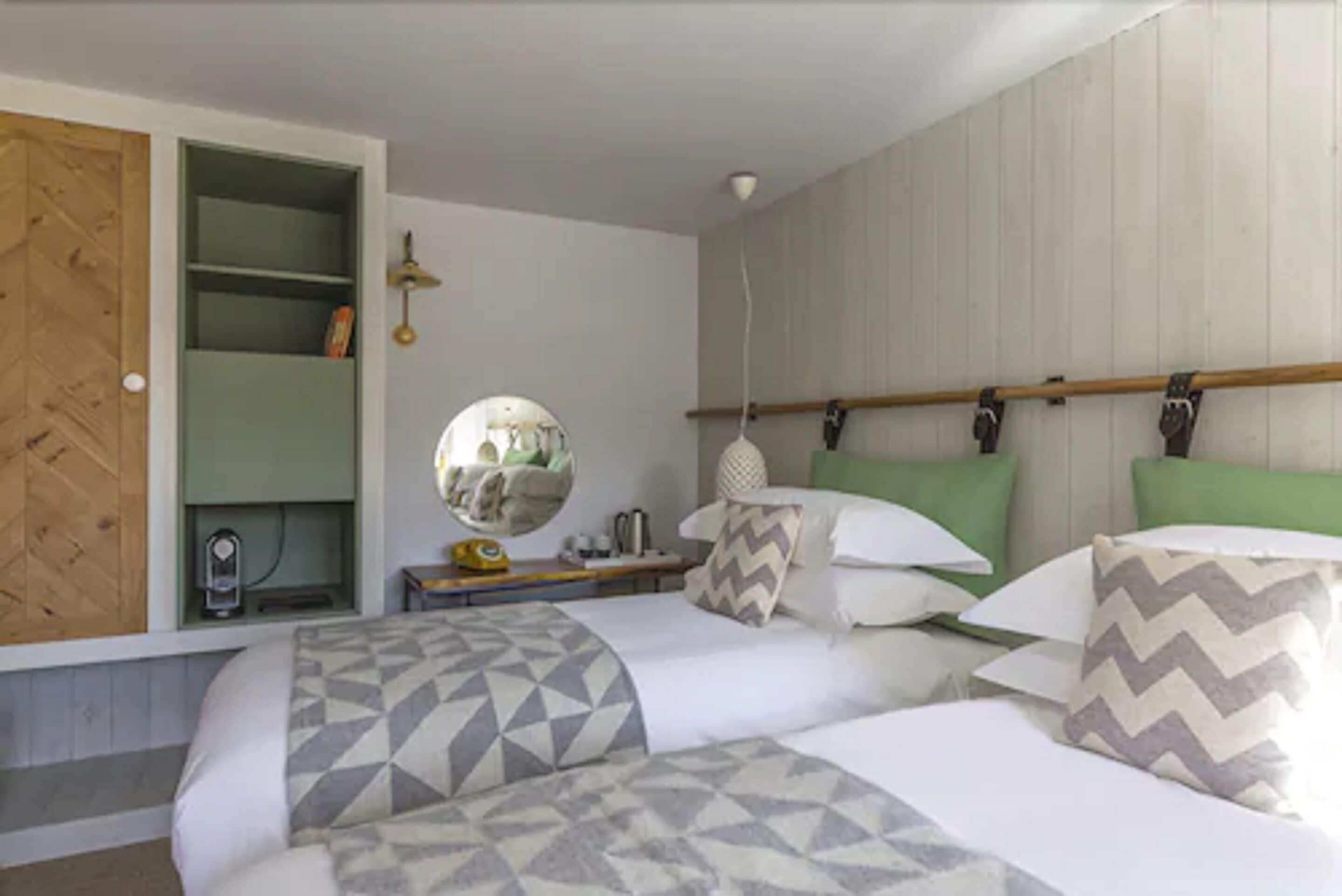 A bedroom with two beds, a wooden wardrobe, a mirror, and a bedside table with a lamp.