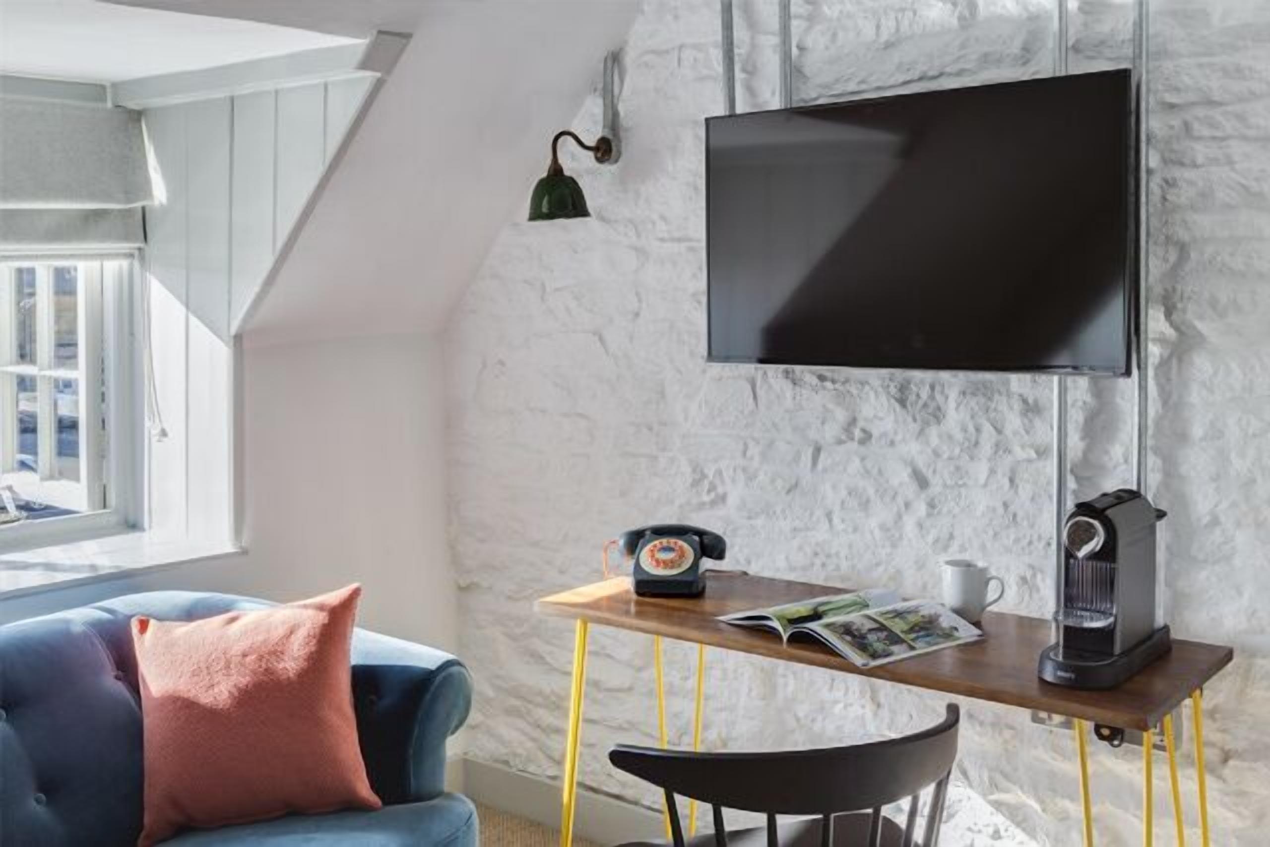 Uma sala de estar moderna com uma TV de tela plana presa a uma parede branca texturizada, uma mesa de centro de madeira com um telefone antigo e uma cafeteira, um sofá azul com uma almofada rosa, e uma janela com vista para edifícios.
