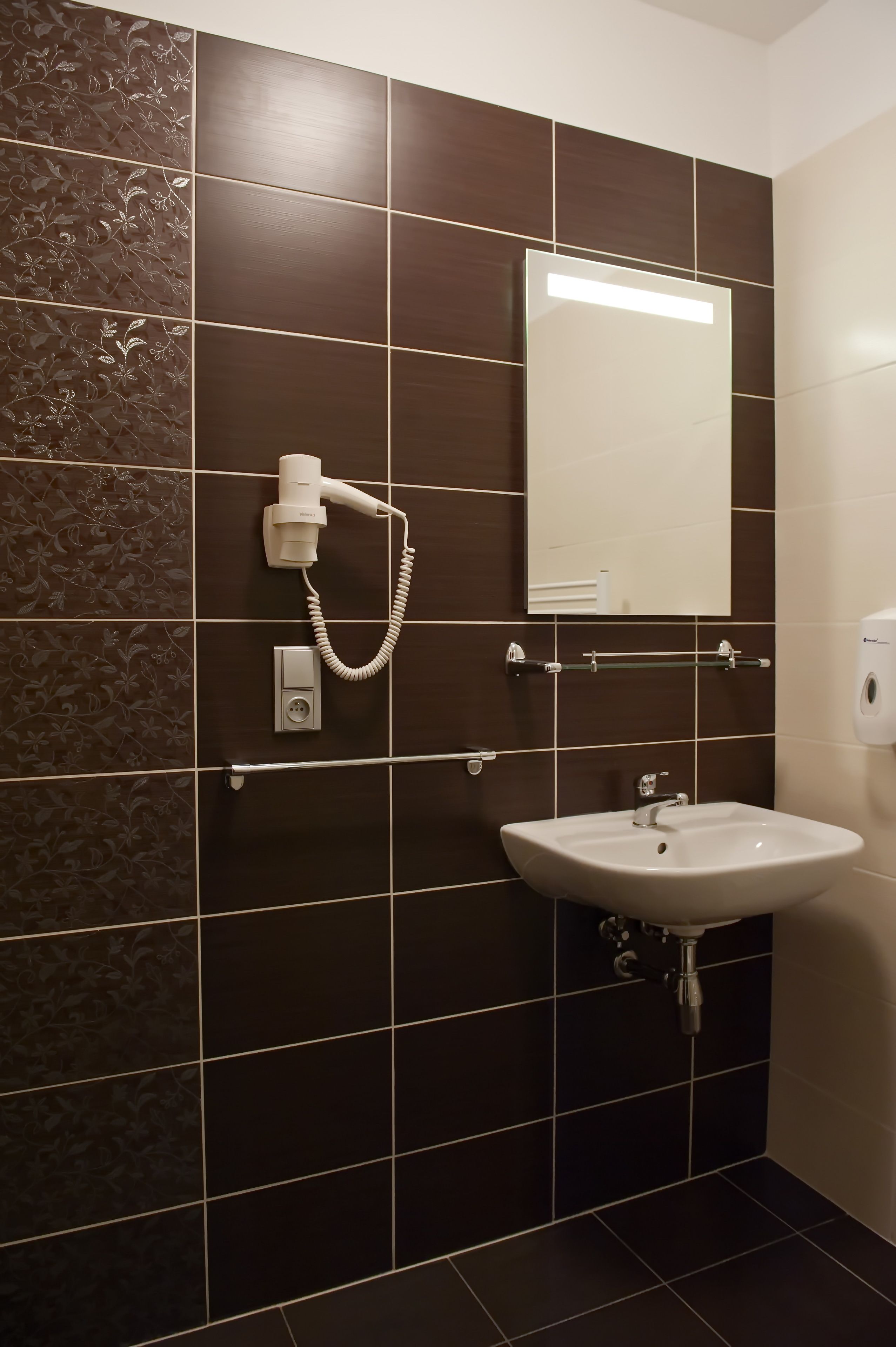 A bathroom with dark brown tiles, a white sink, a mirror, and a hairdryer mounted on the wall.