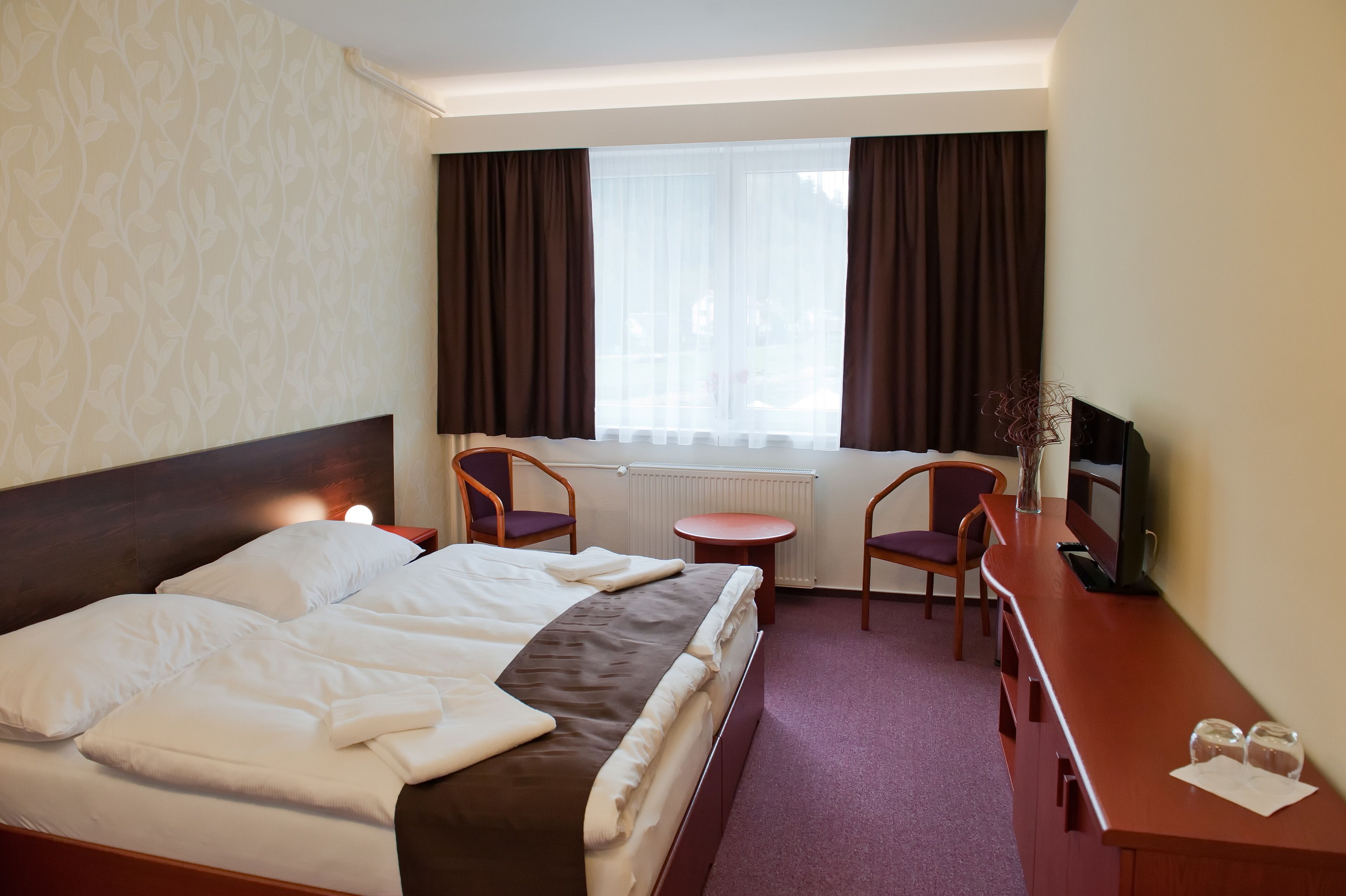 Una habitación de hotel con una cama, dos sillas, una mesita, un televisor y una ventana con cortinas.
