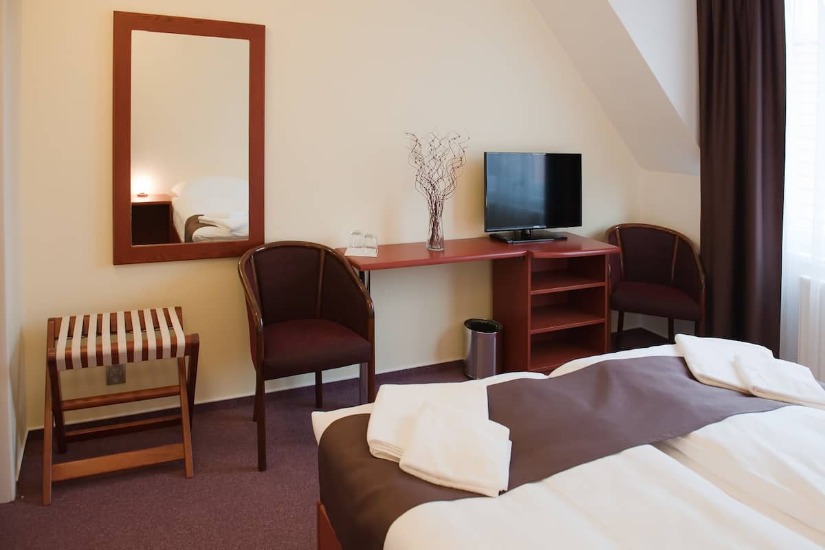 Habitación de hotel con cama, dos sillas, escritorio con televisor, espejo, pequeño banco y cesto de basura.