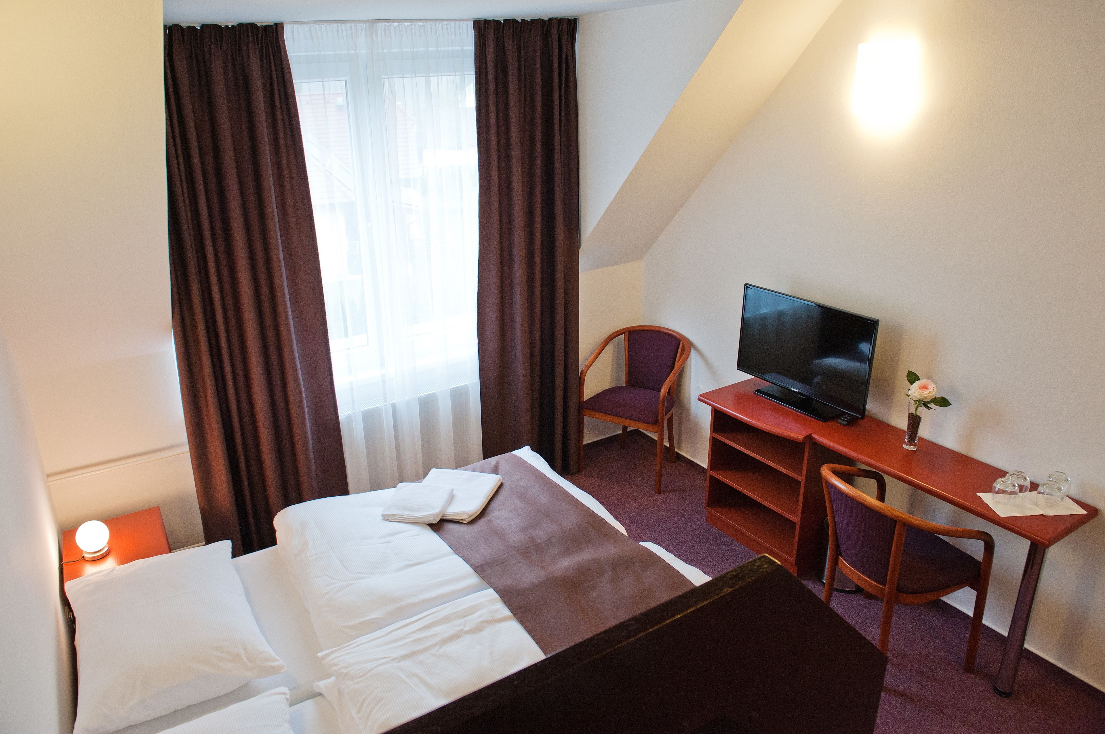 Quarto de hotel com uma cama, uma mesa com televisão, uma cadeira e uma mesa de cabeceira com abajur.