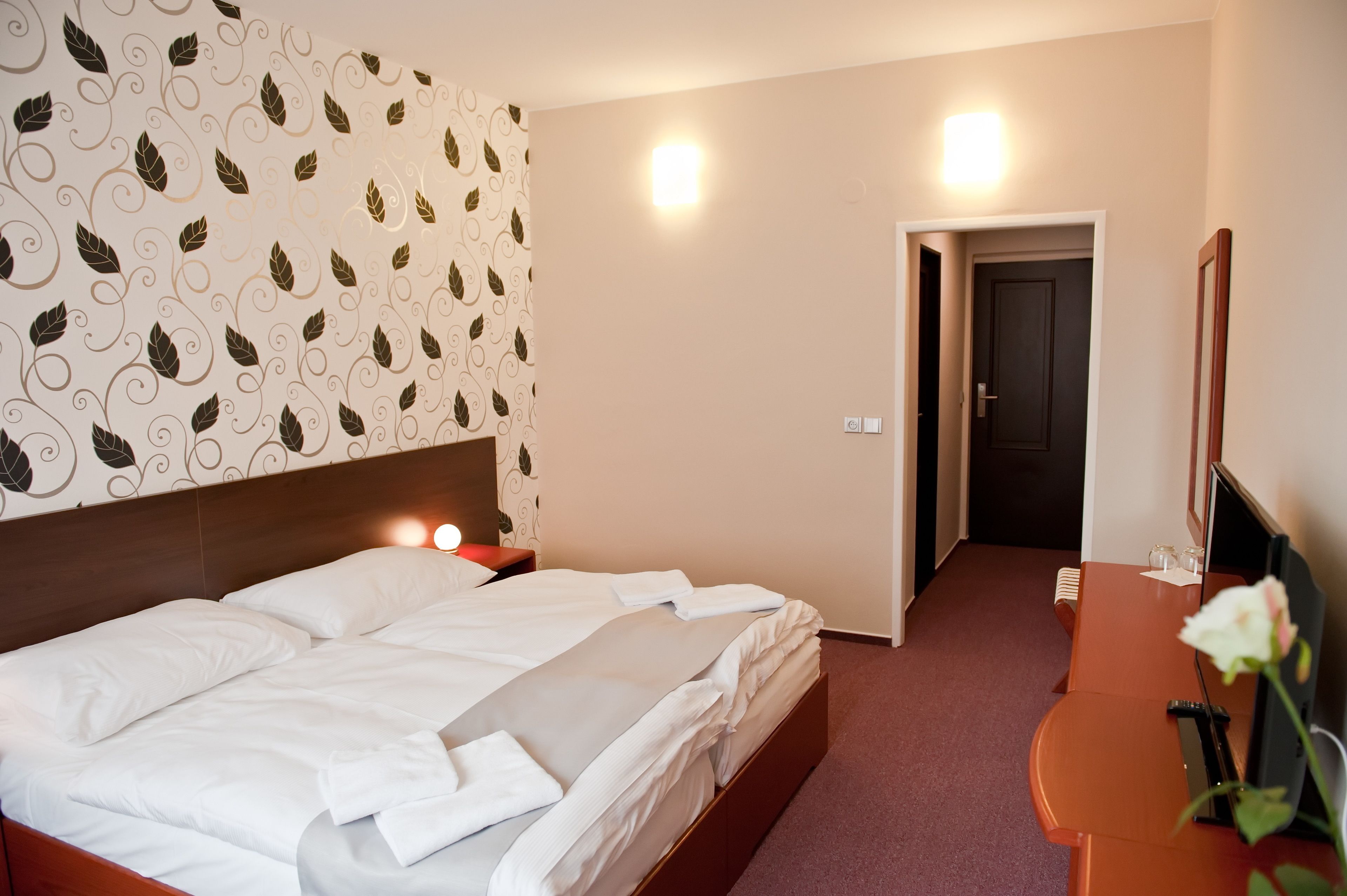 A hotel room with a neatly made bed, a wooden headboard, a bedside table with a lamp, a small table with a vase of flowers, and a television.