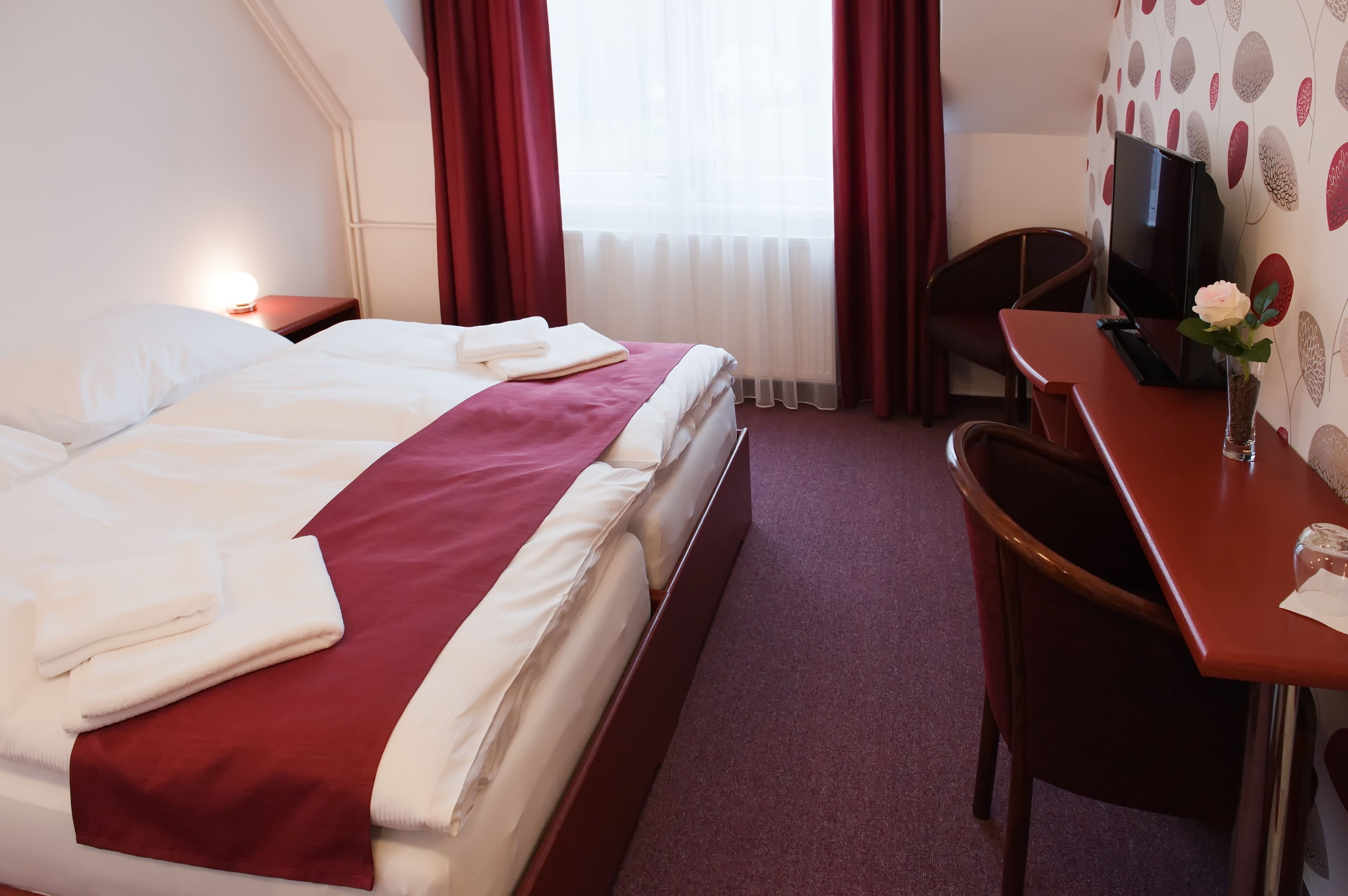 A hotel room with a neatly made bed, a desk with a chair, a television, and a vase with a flower.