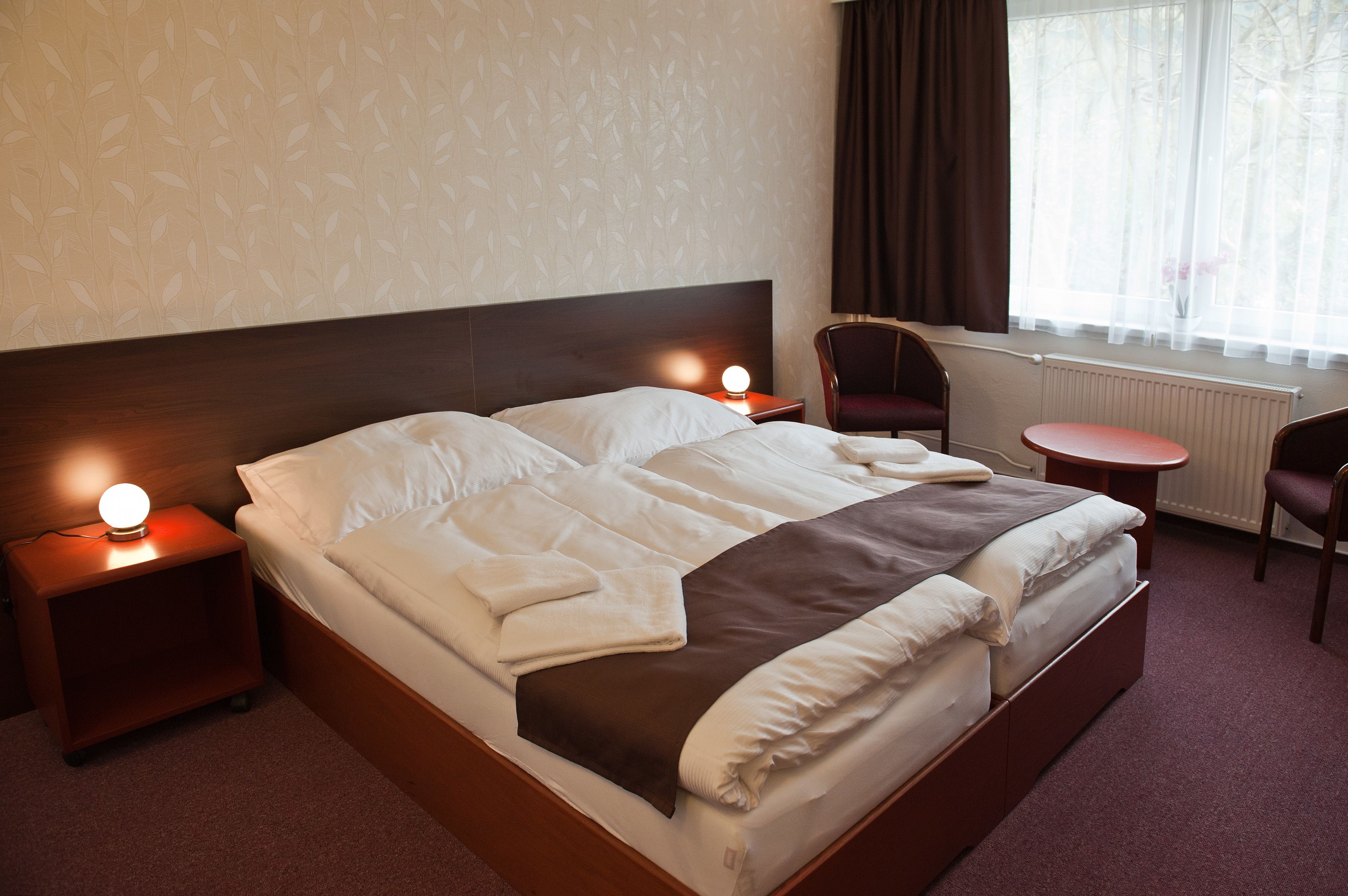 A neatly made bed with white and brown bedding, two bedside tables with lamps, a chair, and a small table.