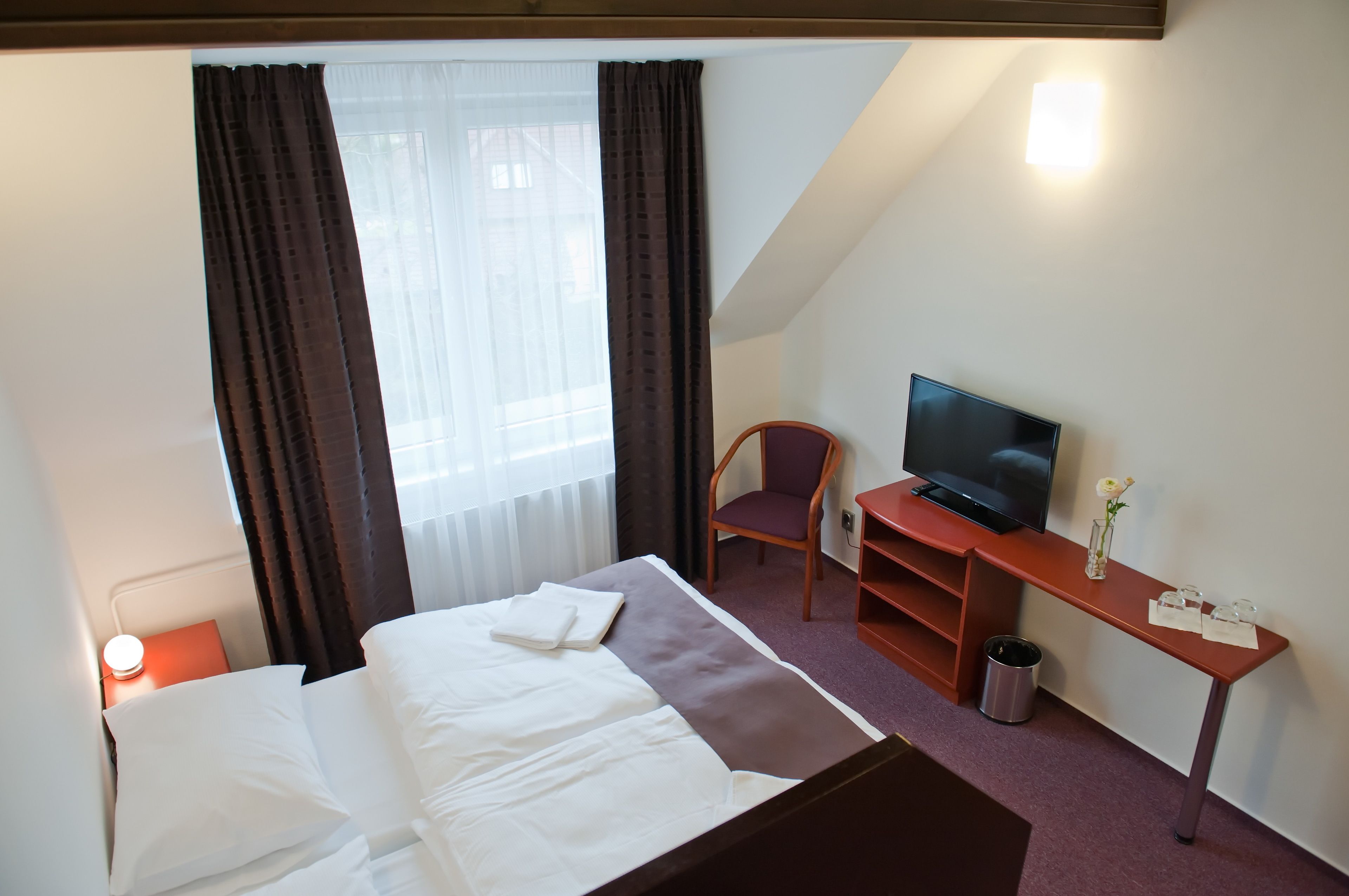A hotel room with a bed, a television on a desk, a chair, and a small table with a vase.