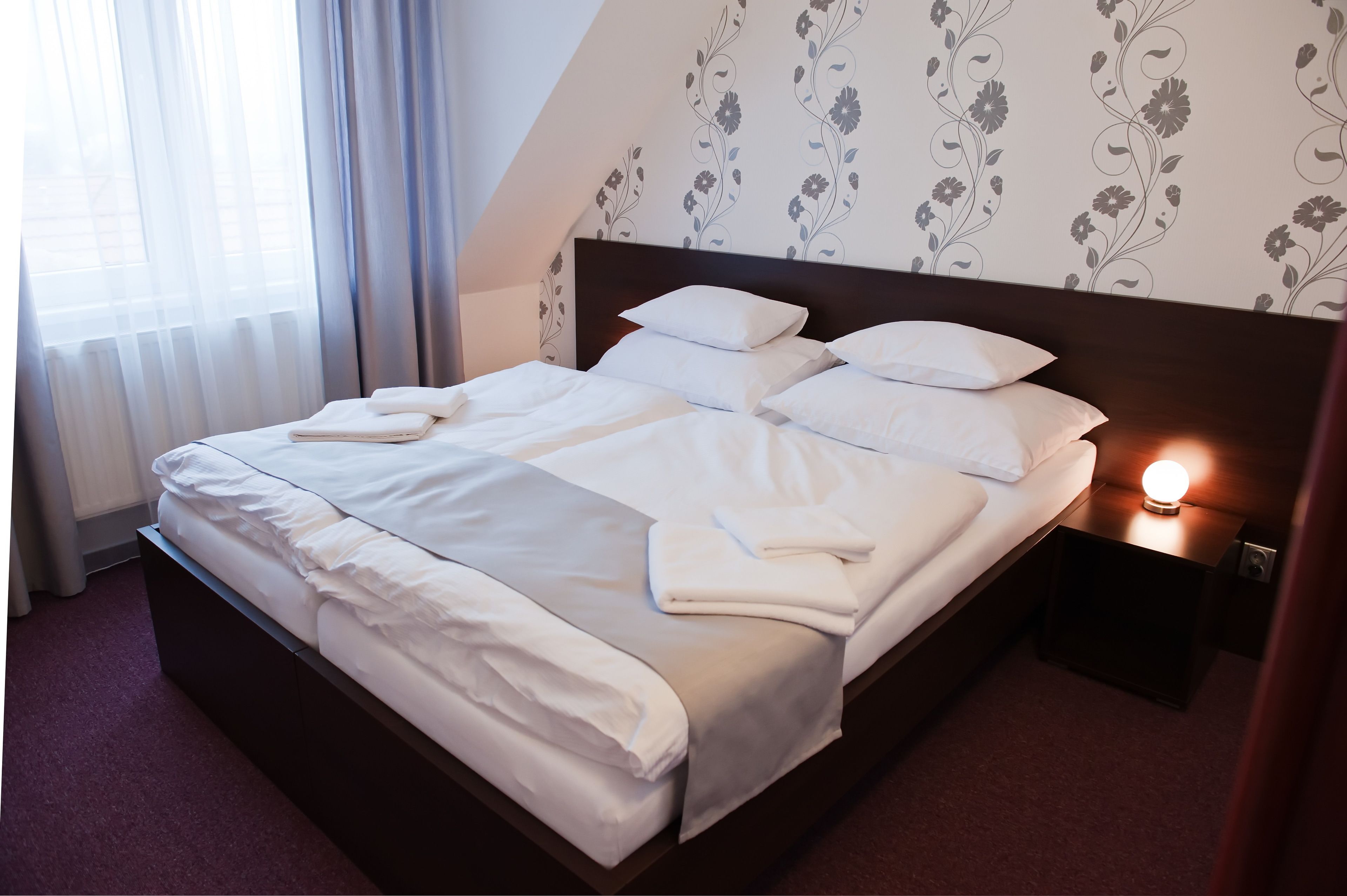 A neatly made bed with white linens and pillows, a bedside table with a lamp, and a floral wallpaper pattern on the wall.