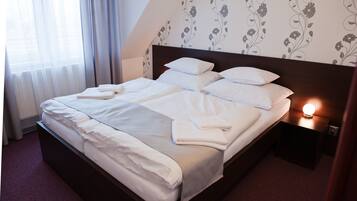A neatly made bed with white linens and pillows, a bedside table with a lamp, and a floral wallpaper pattern on the wall.