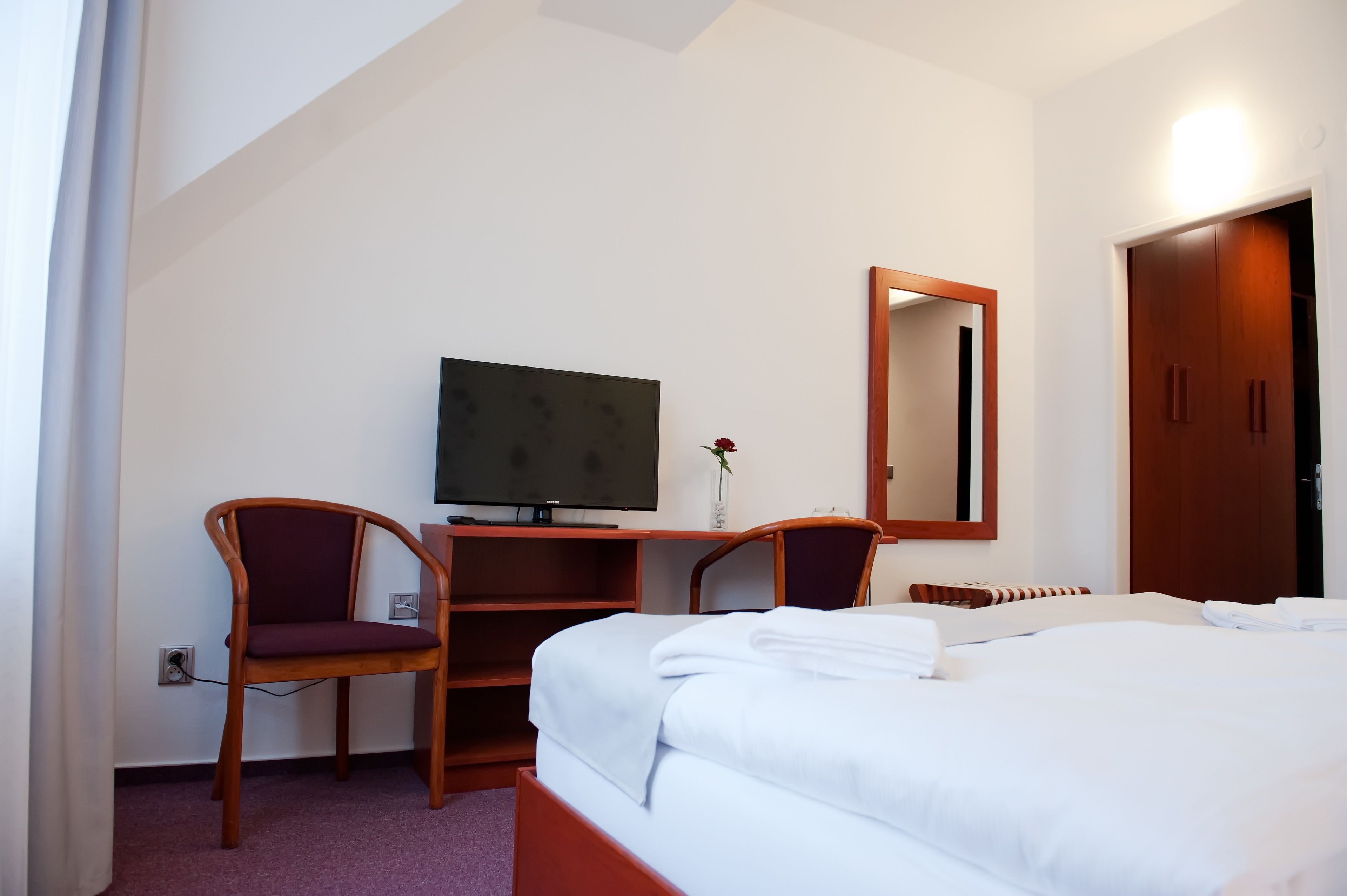 A hotel room with a bed, a desk with a television, two chairs, and a mirror.