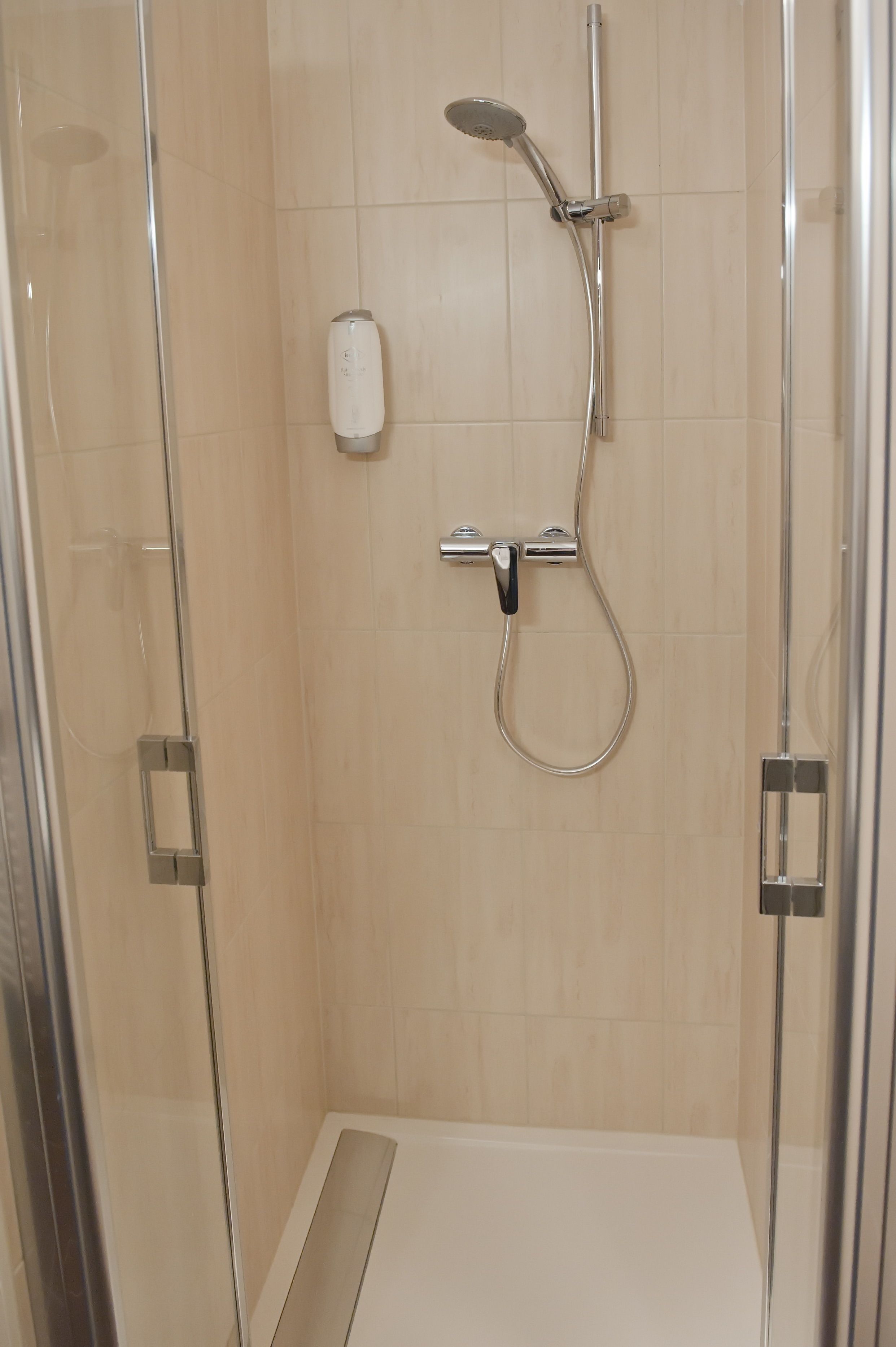A glass-enclosed shower stall with a handheld showerhead, a wall-mounted faucet, and a soap dispenser.