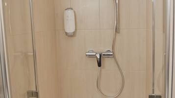 A glass-enclosed shower stall with a handheld showerhead, a wall-mounted faucet, and a soap dispenser.