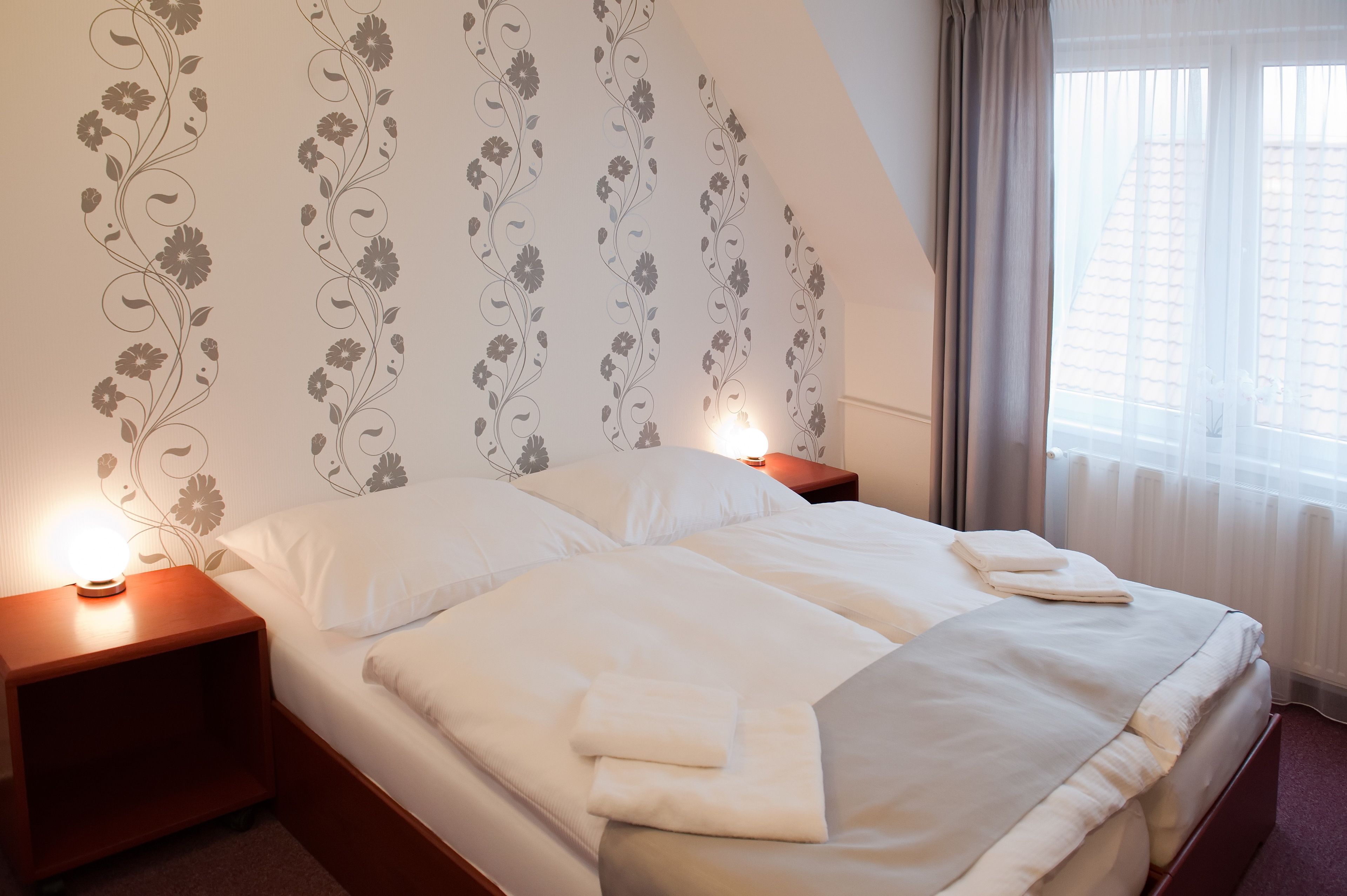 Una cama bien hecha con ropa blanca y toallas, una mesita de noche de madera con una lámpara, y un diseño de papel tapiz floral en la pared.