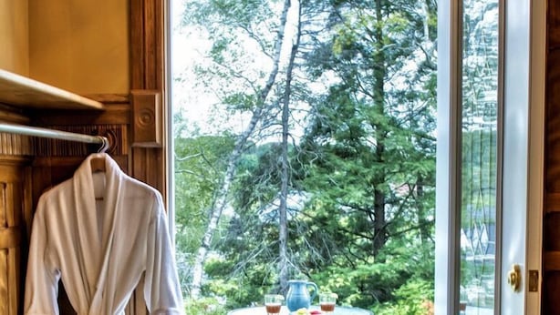 A wooden interior with a white bathrobe hanging on a rack, a round table with chairs outside, and a view of a forest through a glass door. - Shamrock Suites (Florenceville-Bristol)