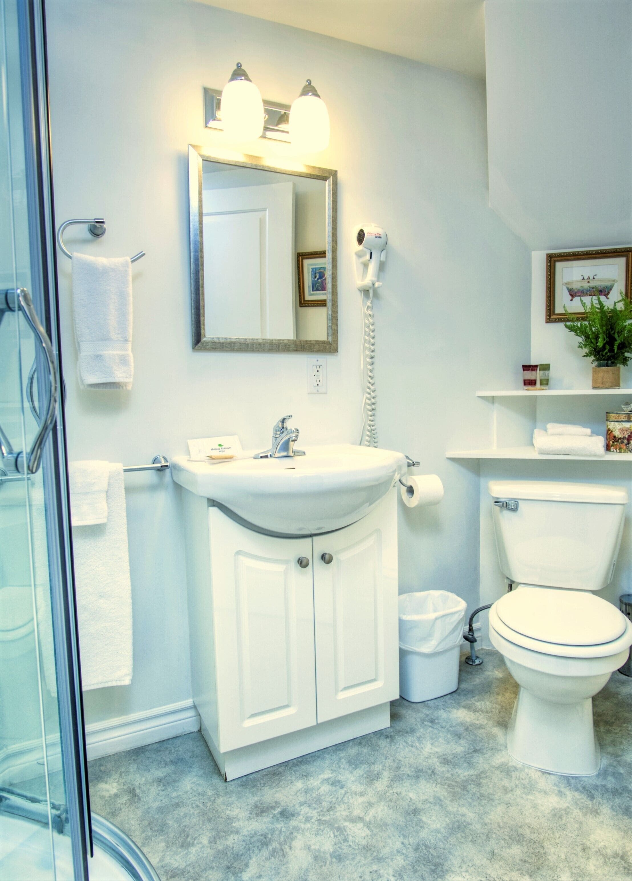 Une salle de bain avec un lavabo blanc, un miroir et des toilettes. On y trouve un sèche-cheveux fixé au mur, un porte-serviettes et une poubelle.