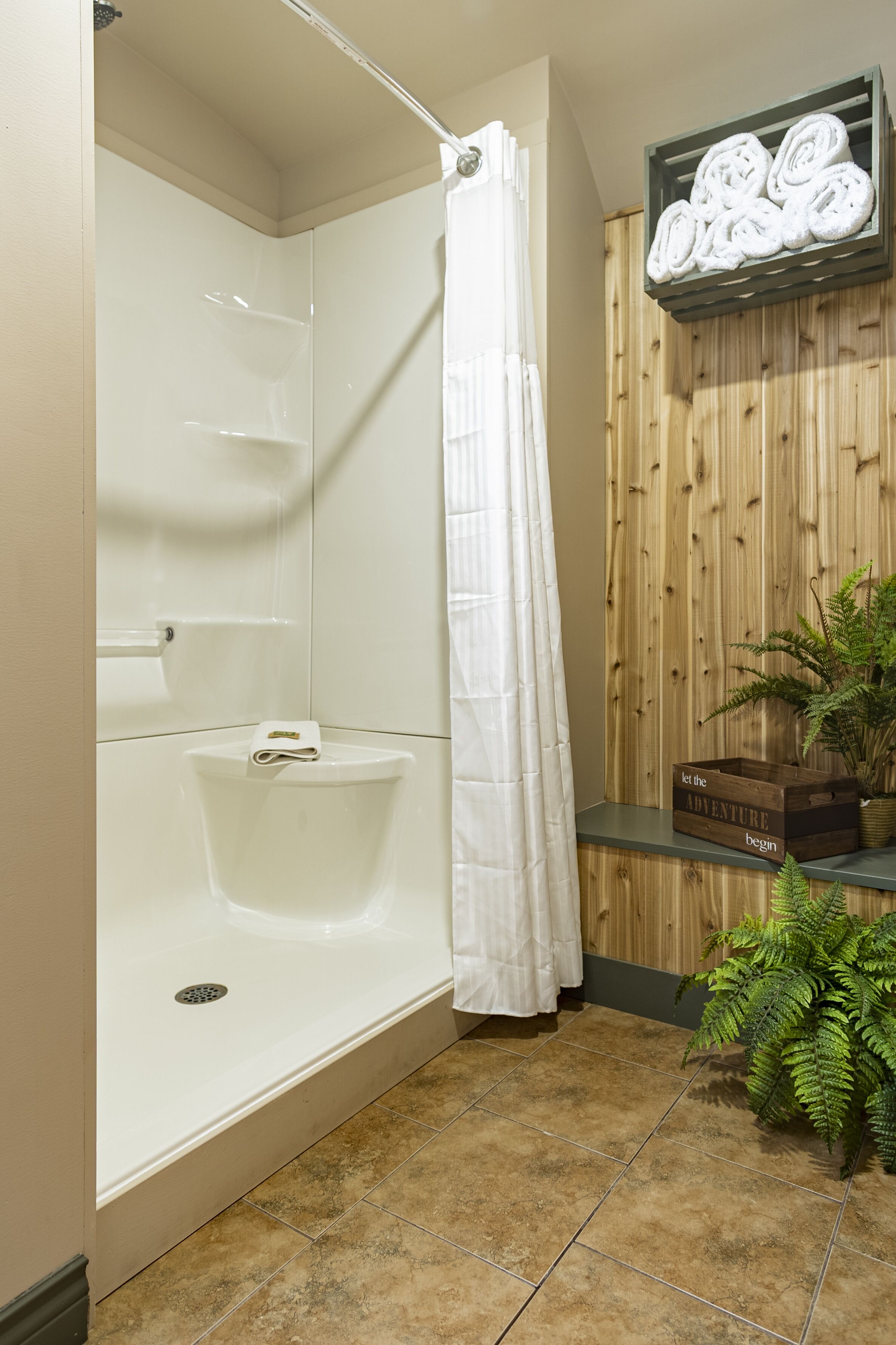 Baño con una bañera blanca, cortina de ducha, pared de paneles de madera, estante con toallas enrolladas y una planta en maceta.