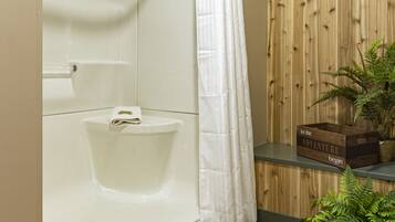 Baño con una bañera blanca, cortina de ducha, pared de paneles de madera, estante con toallas enrolladas y una planta en maceta.