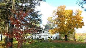 A white house with a symmetrical front, surrounded by a well-maintained lawn and trees with autumn foliage. - Shamrock Suites (Florenceville-Bristol)