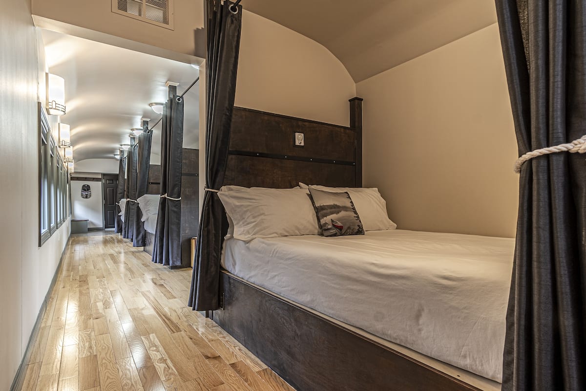 Ein schmaler Korridor mit Holzboden führt zu einem Raum mit einem Einzelbett, einem dunklen Kopfteil und Vorhängen.